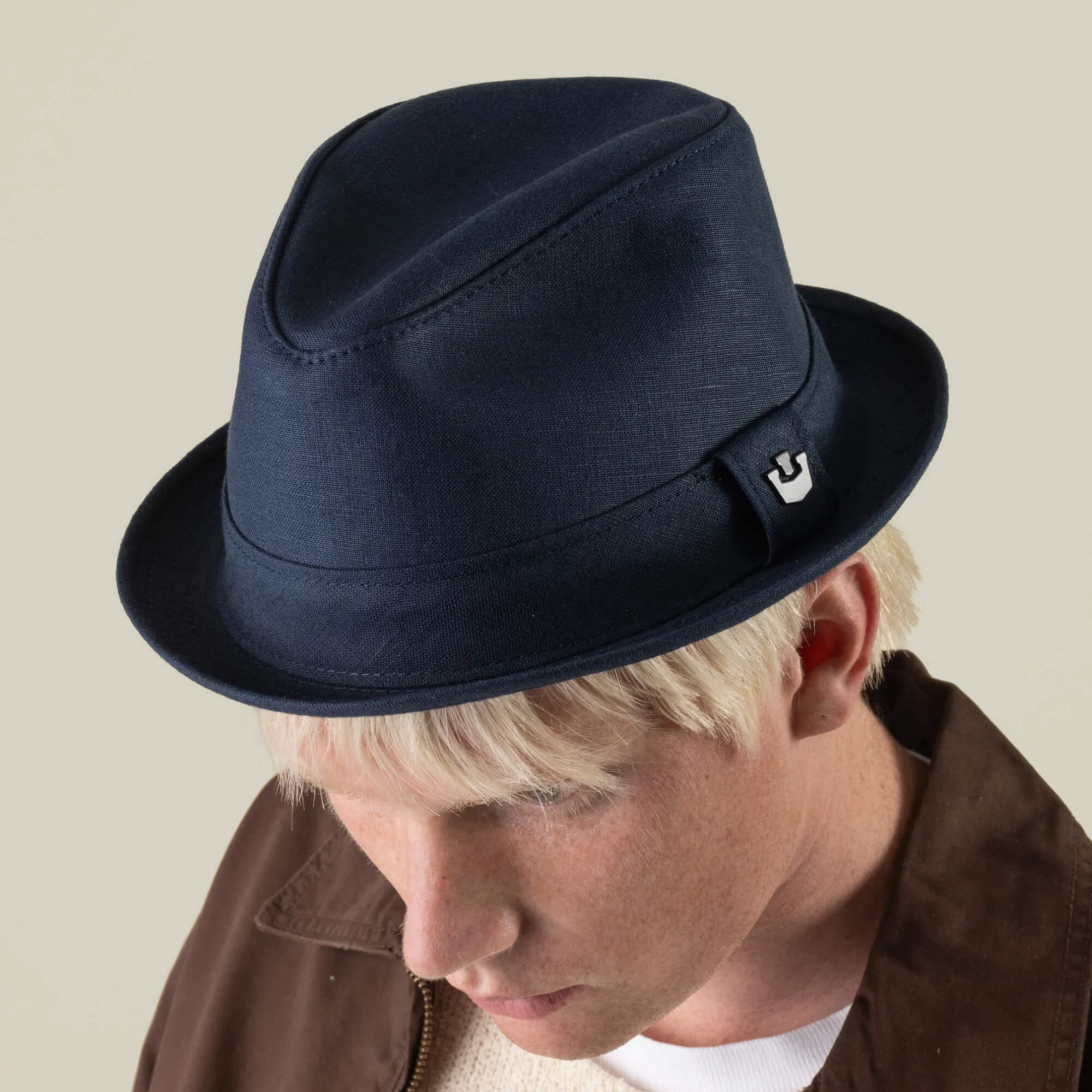 A person with blond hair, dressed in a brown jacket and Charlie Pants by Goorin Bros., wearing an edge navy fedora and looking downward against a plain background. The linen fedora is hand-cut in the USA.