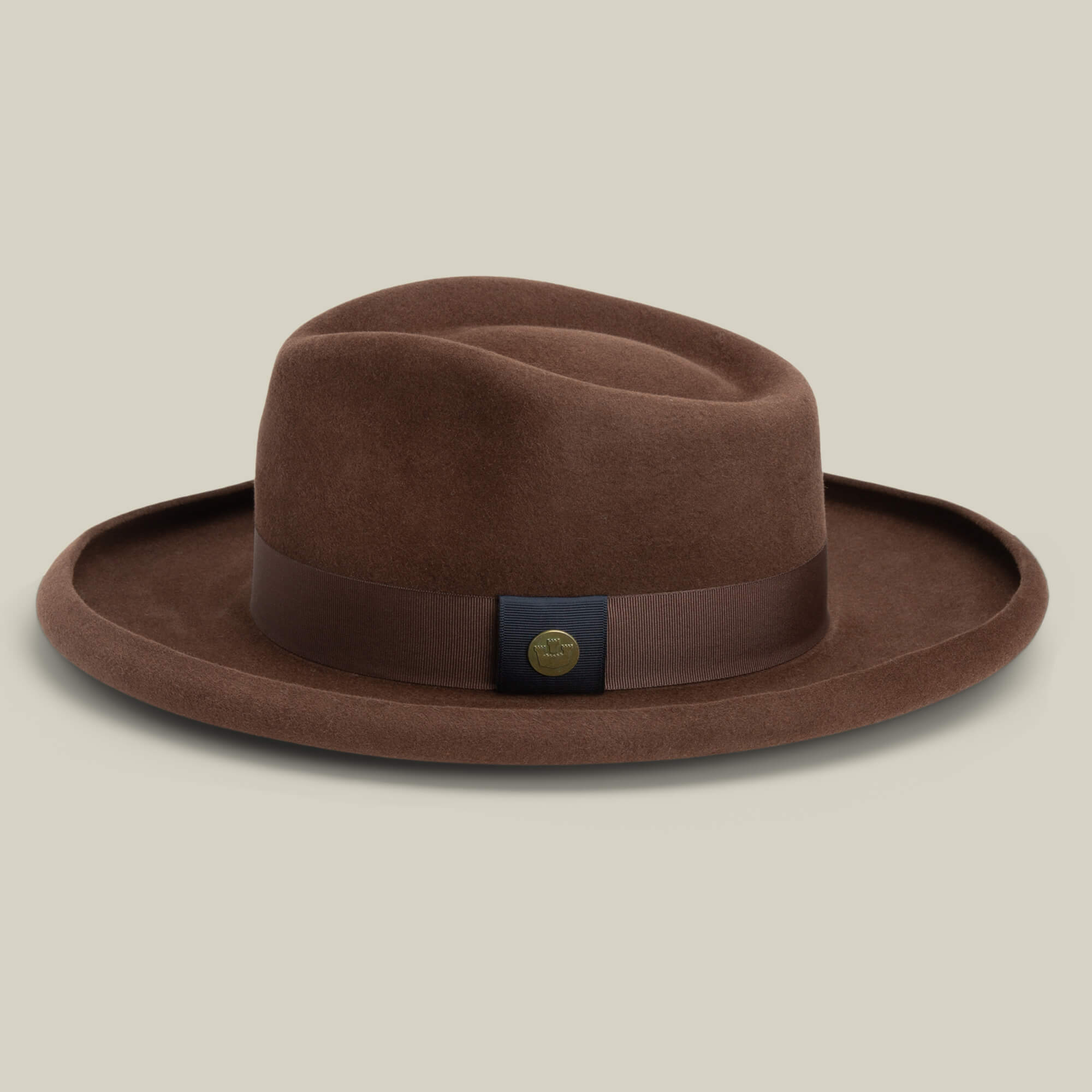 The Goorin Bros. Colonel Pierce is a brown hat with a pencil curl brim, matching band, and round metallic emblem, displayed on a plain beige background.