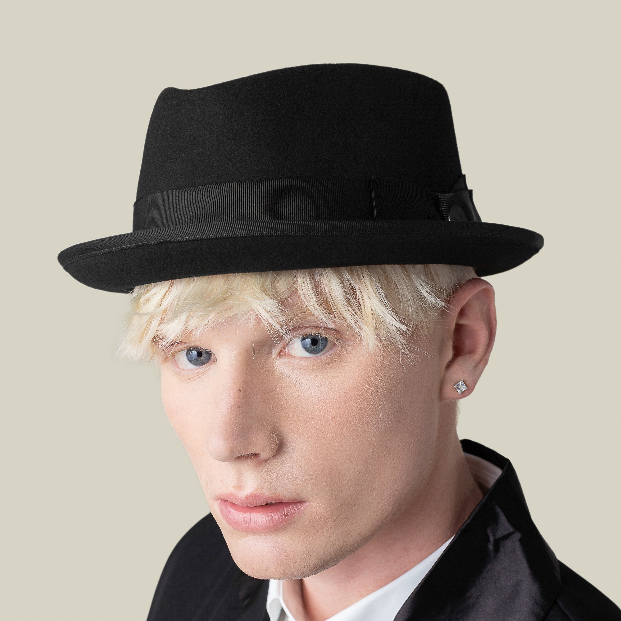 A person with light skin, blonde hair, and blue eyes wears the Slick Carter black wool short brim fedora by Goorin Bros., paired with a black blazer and stud earring, looking stylishly at the camera against a beige background.