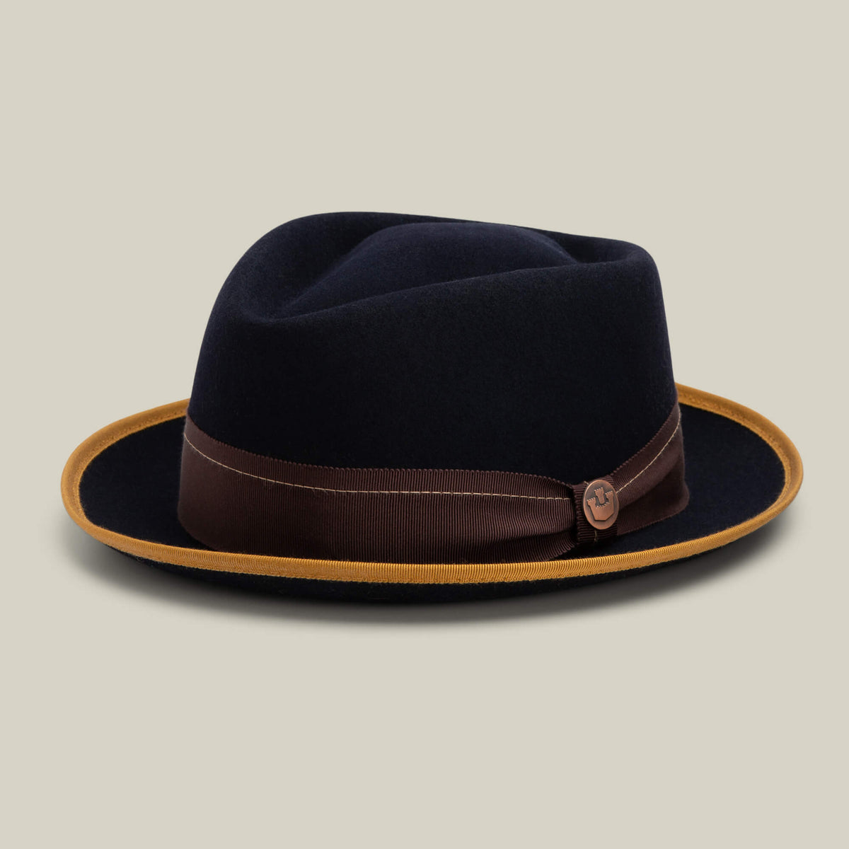 The Goorin Bros. Slick Carter is a dark wool short-brim fedora featuring a brown ribbon band accented with a small emblem and a contrasting mustard-colored edge, set against a plain beige background—a modern tribute to classic Slick Carter style.