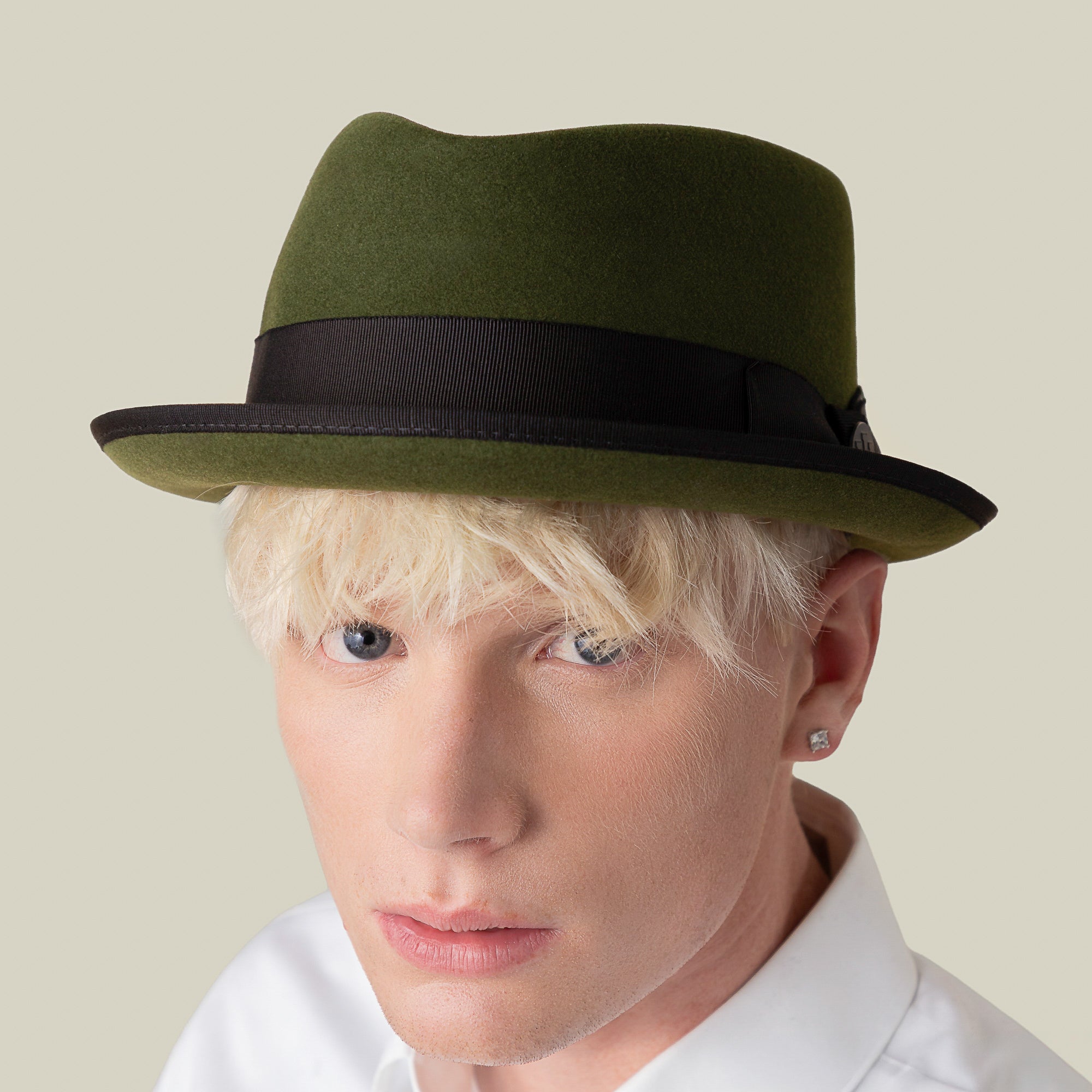 Effortlessly stylish, a platinum blonde person wears the Goorin Bros. Slick Carter green wool short brim fedora with a black band, paired with a white collared shirt and diamond stud earring, facing the camera against a plain background.