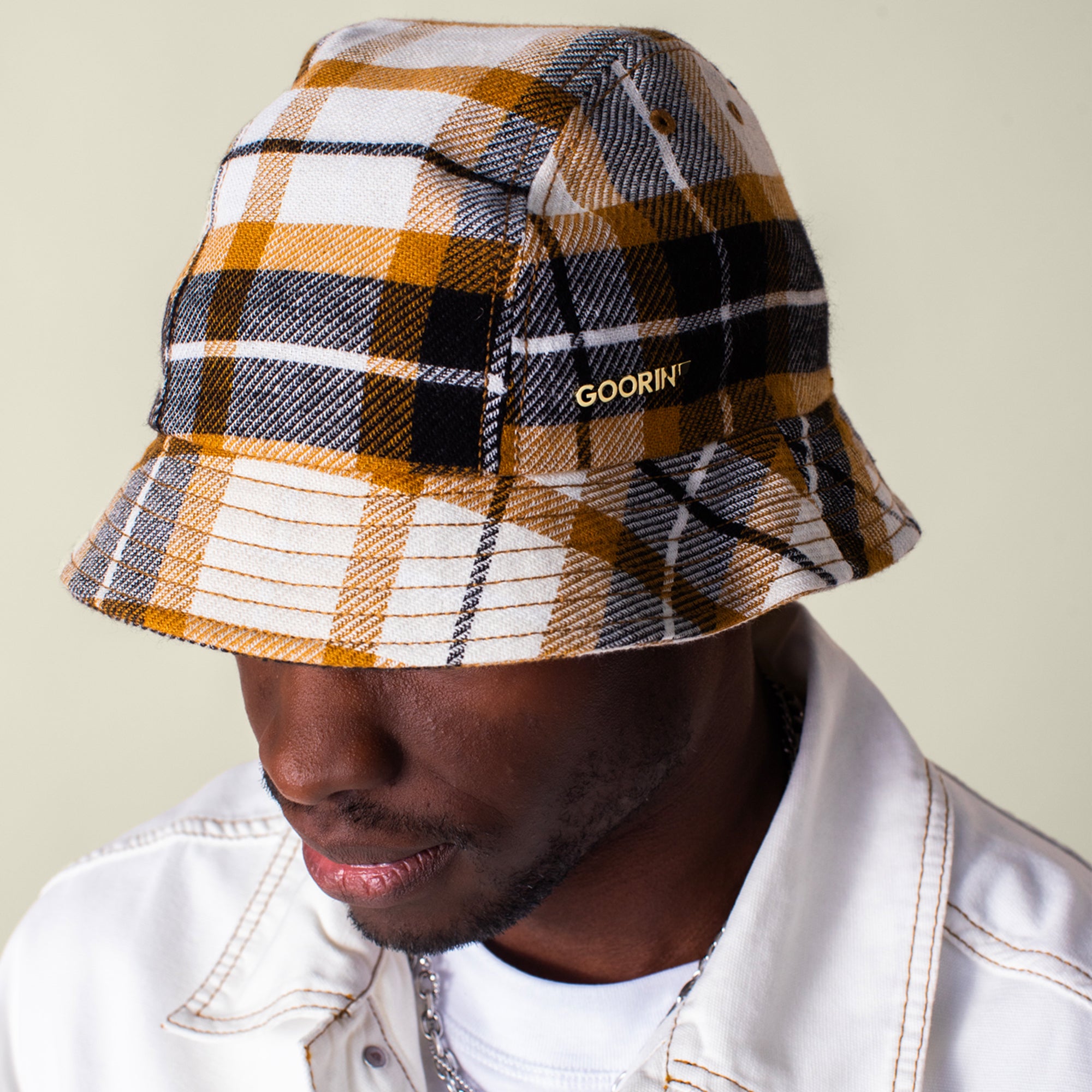 A person wears a white jacket and the Bespoke Wordmark Bucket by Goorin Bros., featuring a yellow, black, and white plaid design with an embroidered GOORIN wordmark on the side.