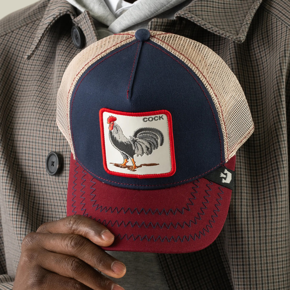 A person holds The Farm’s “The Cock” baseball cap in navy and burgundy, featuring a rooster patch labeled COCK. They are dressed in a plaid jacket and gray hoodie.