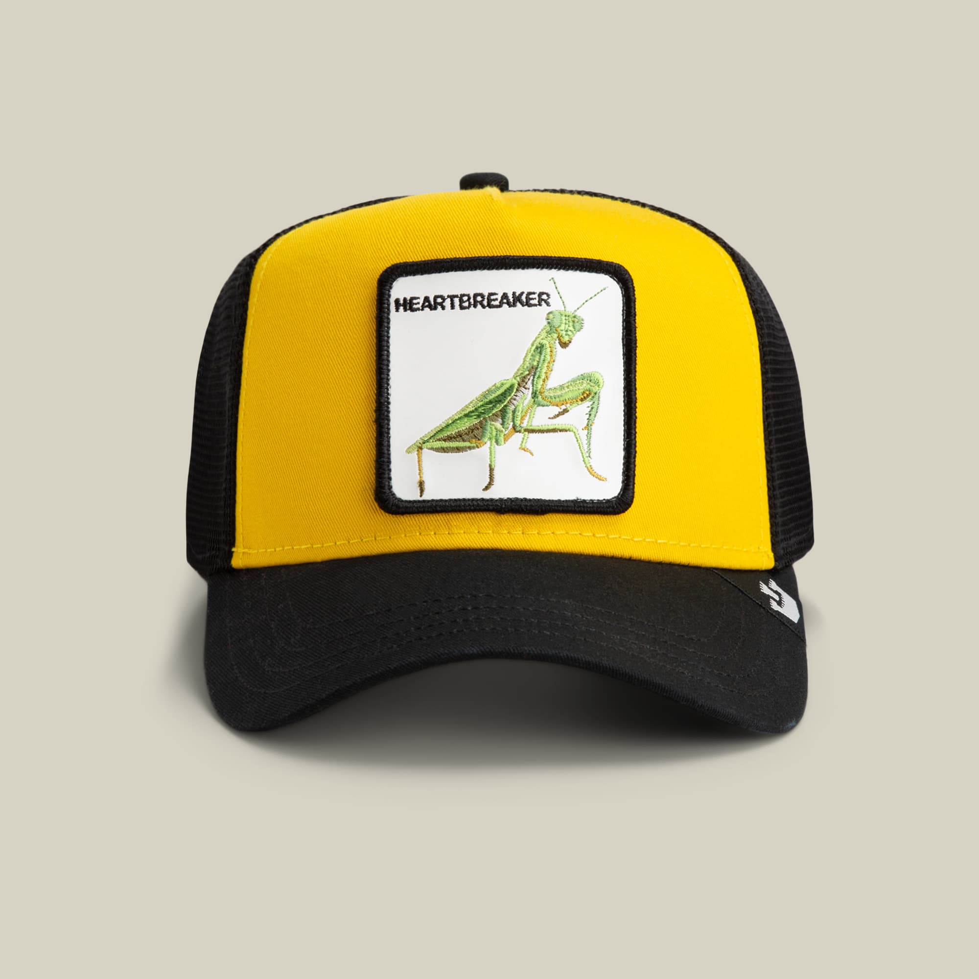 The Heartbreaker by Goorin Bros. is a yellow and black mesh trucker hat featuring a embroidered patch of a green praying mantis insect with the word "Heartbreaker" text, shown on a neutral studio background.