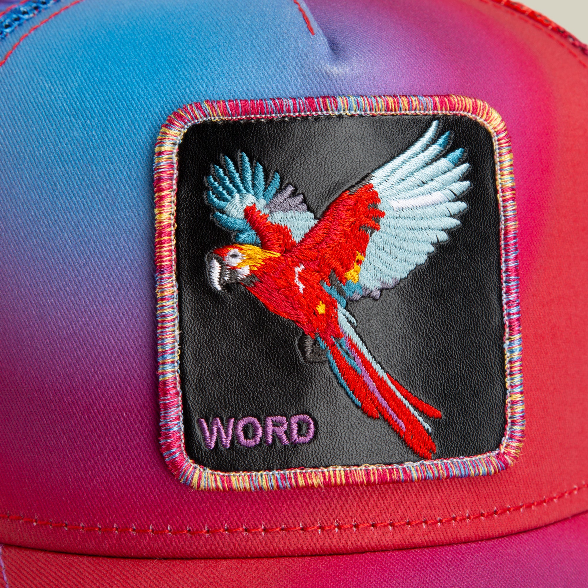 Close-up of The Farm’s “Bird is the Word” cap, showcasing a red parrot in flight and the word “WORD” stitched in purple on a black patch. All Sales Are Final.
