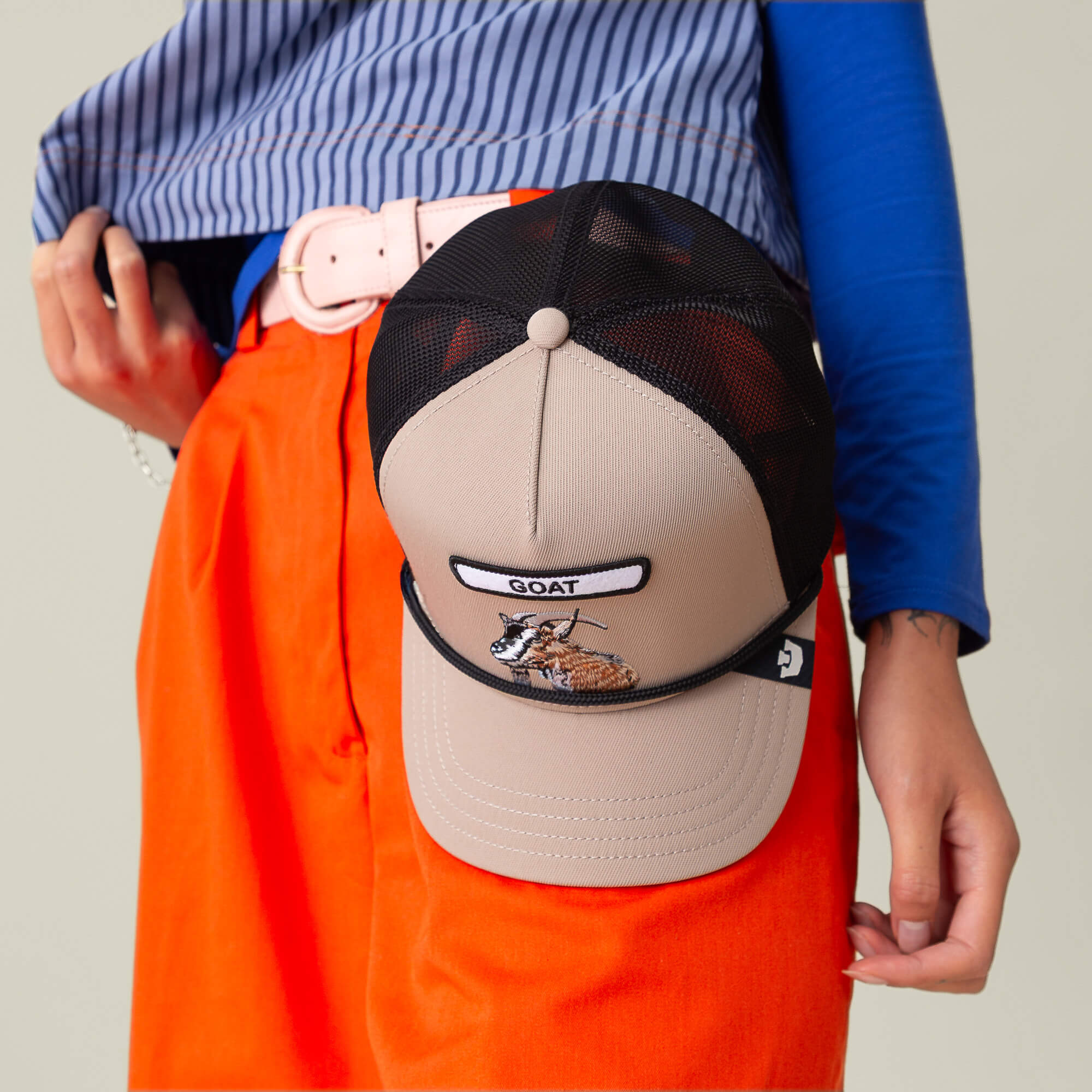 A person in orange pants and a blue shirt holds The Farm's GB2 Goat Trucker Hat, a beige and black hat featuring "GOAT" and an embroidered goat design.