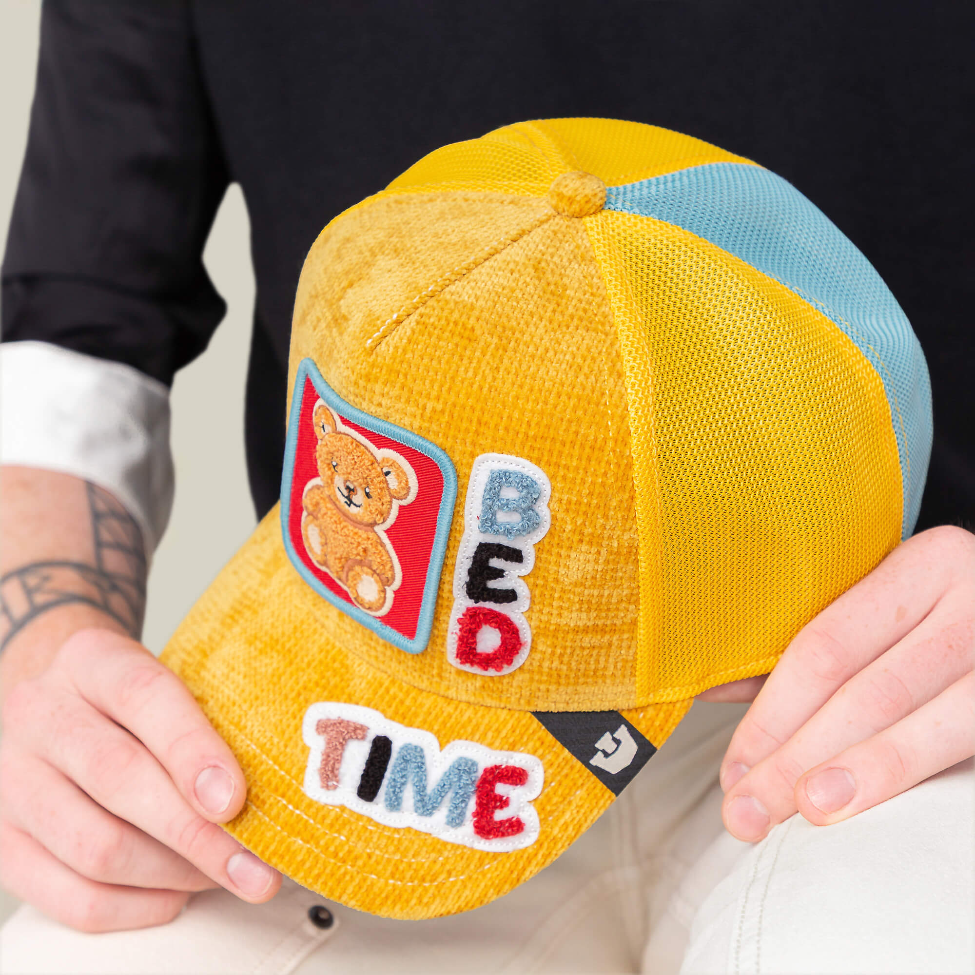 Someone holds a sunshine yellow Goorin Bros. Duvet All Det hat featuring colorful BED TIME and Bed Time Energy patches and a teddy bear illustration on the front.