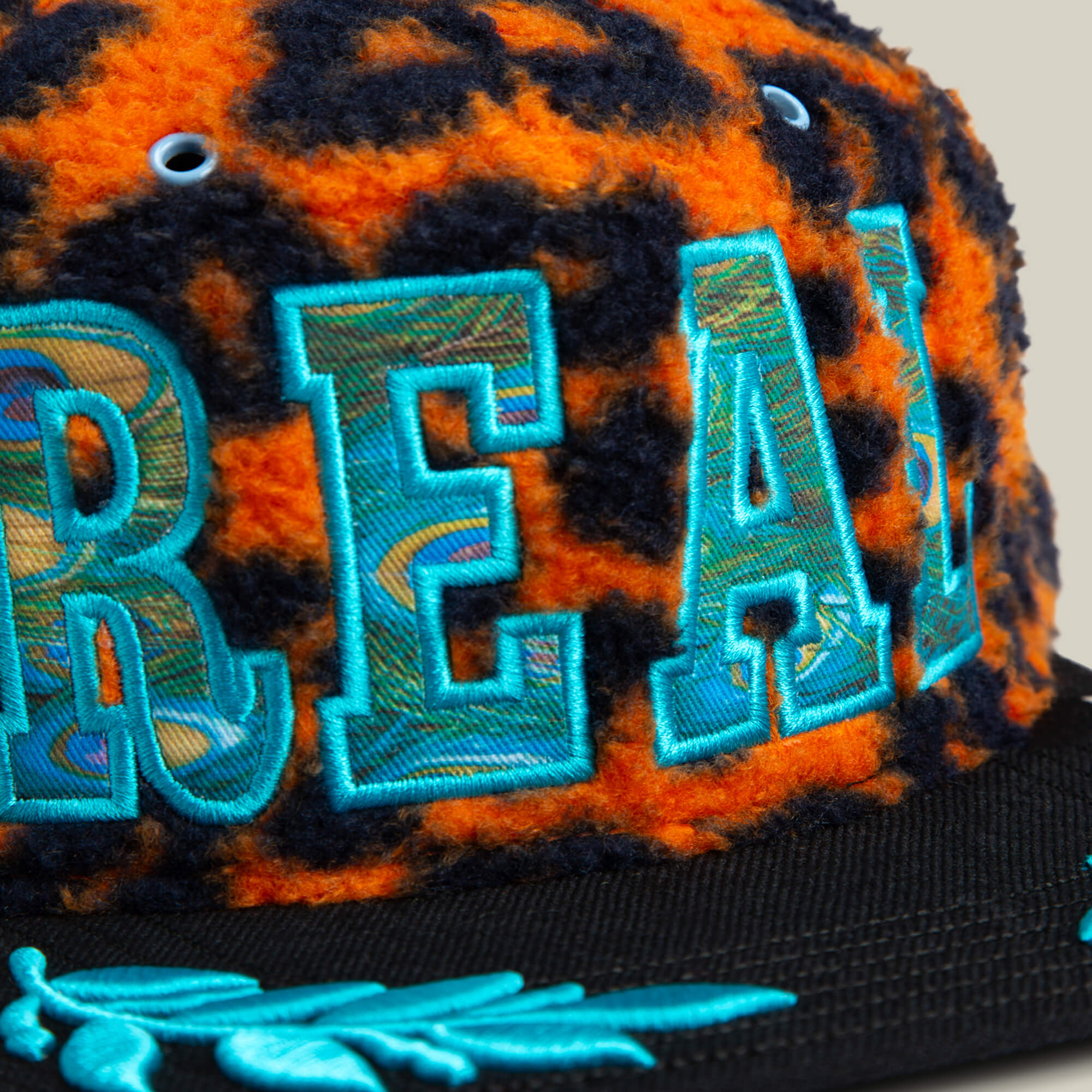 A close-up of the El Camino cap by Goorin Bros. in orange leopard sherpa with blue embroidered letters, a black brim, and an aqua embroidered leaf design.