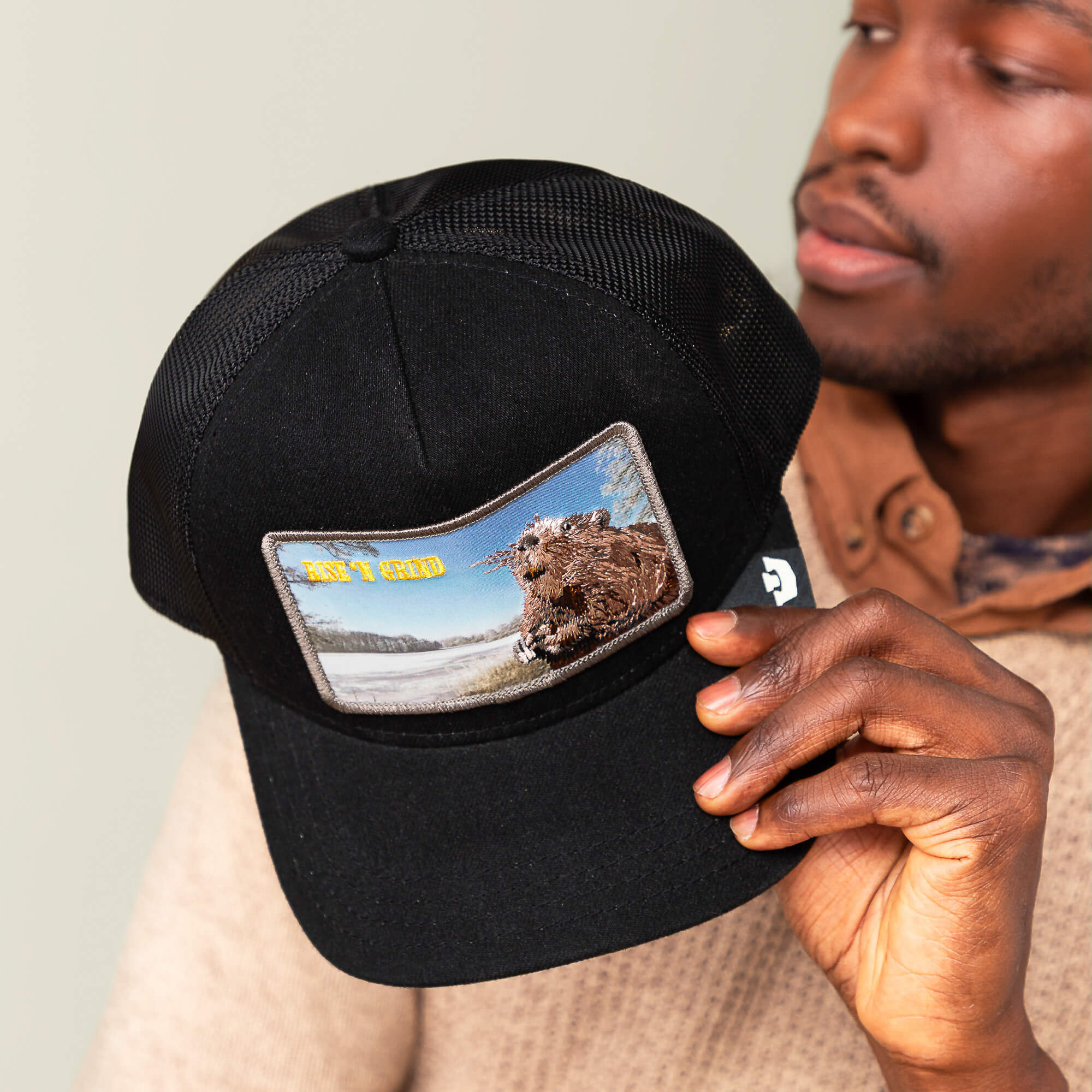 A person holds a black mesh trucker hat by Goorin Bros. called "Morning Wood," featuring a patch with a bear by water and yellow text reading GET Y GEAR—perfect for that rise ’n grind energy.