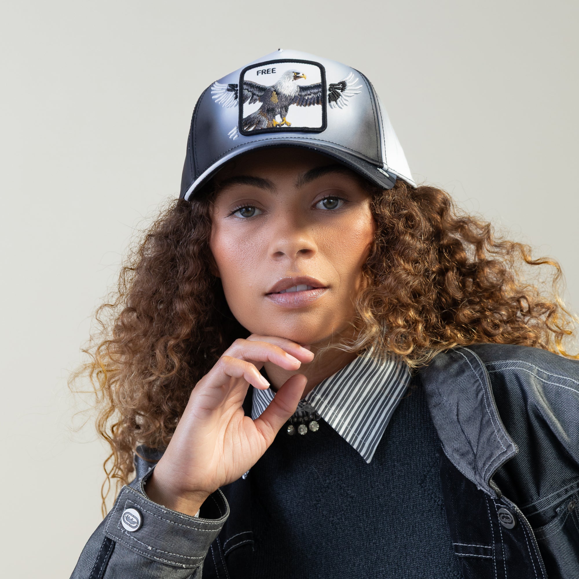 A woman with curly hair wears a black-and-white jacket and the Goorin Bros. Steal This Hat, which features a bold bald eagle patch and the word FREE on the front, set against a plain background.