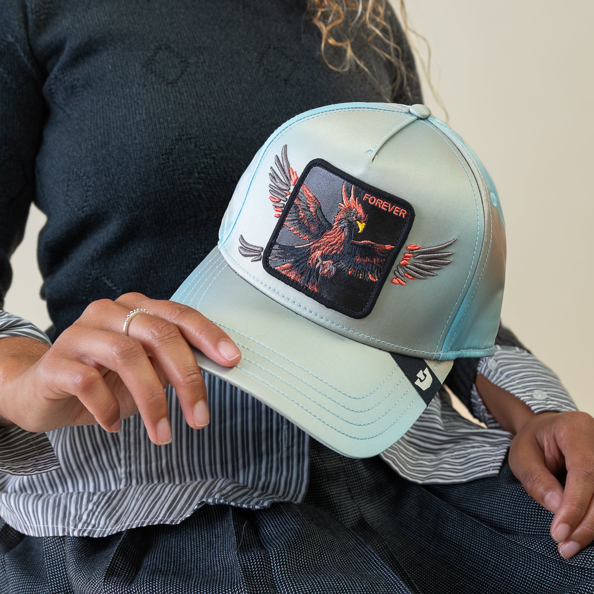 A person holds a light blue Goorin Bros. cap, "Never Stop Never Stopping," featuring a sky-blue crown and an embroidered eagle patch with the word FOREVER above it.