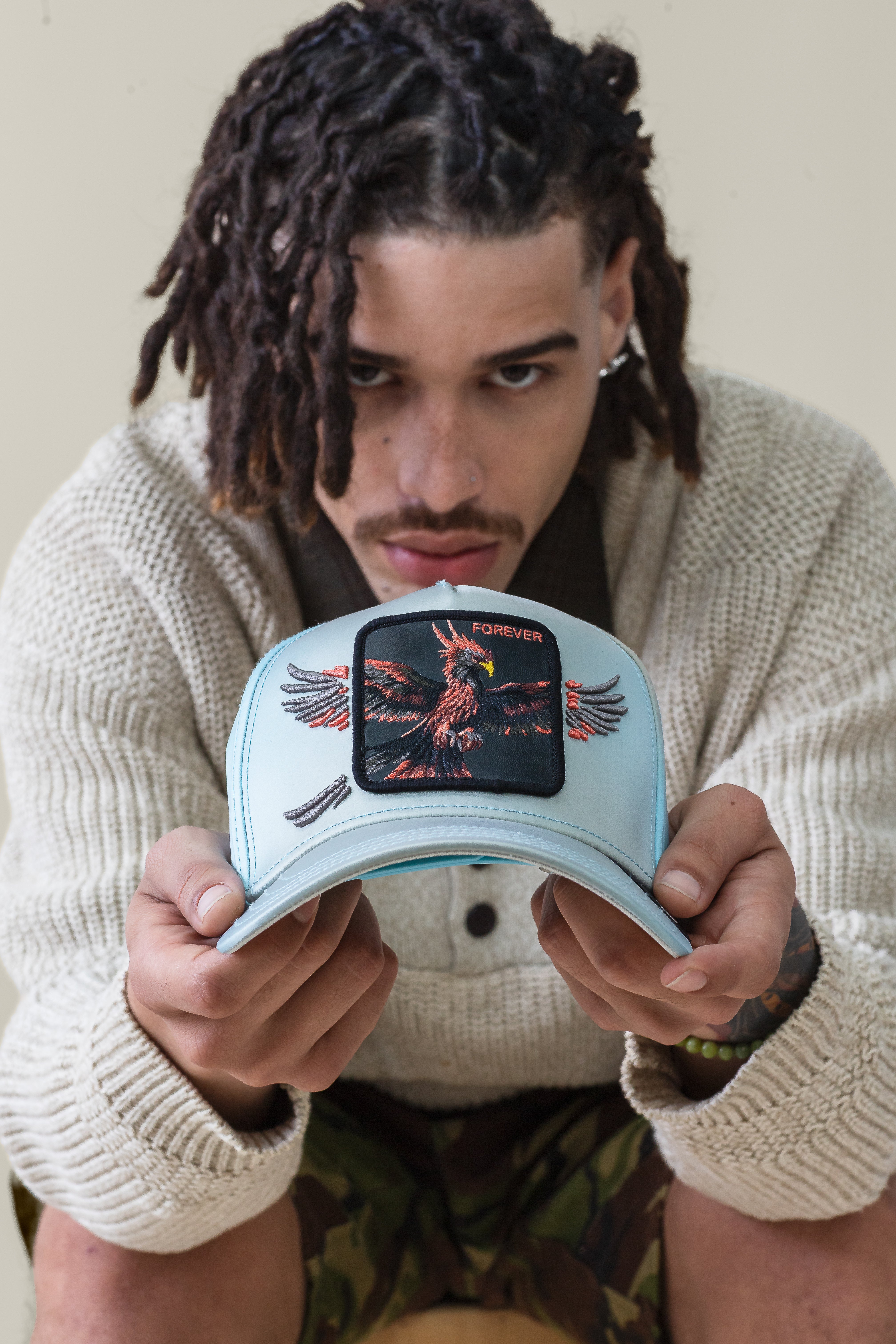 A person with dreadlocks holds out a sky-blue Goorin Bros. "Never Stop Never Stopping" crown featuring an eagle patch with FOREVER embroidered above.