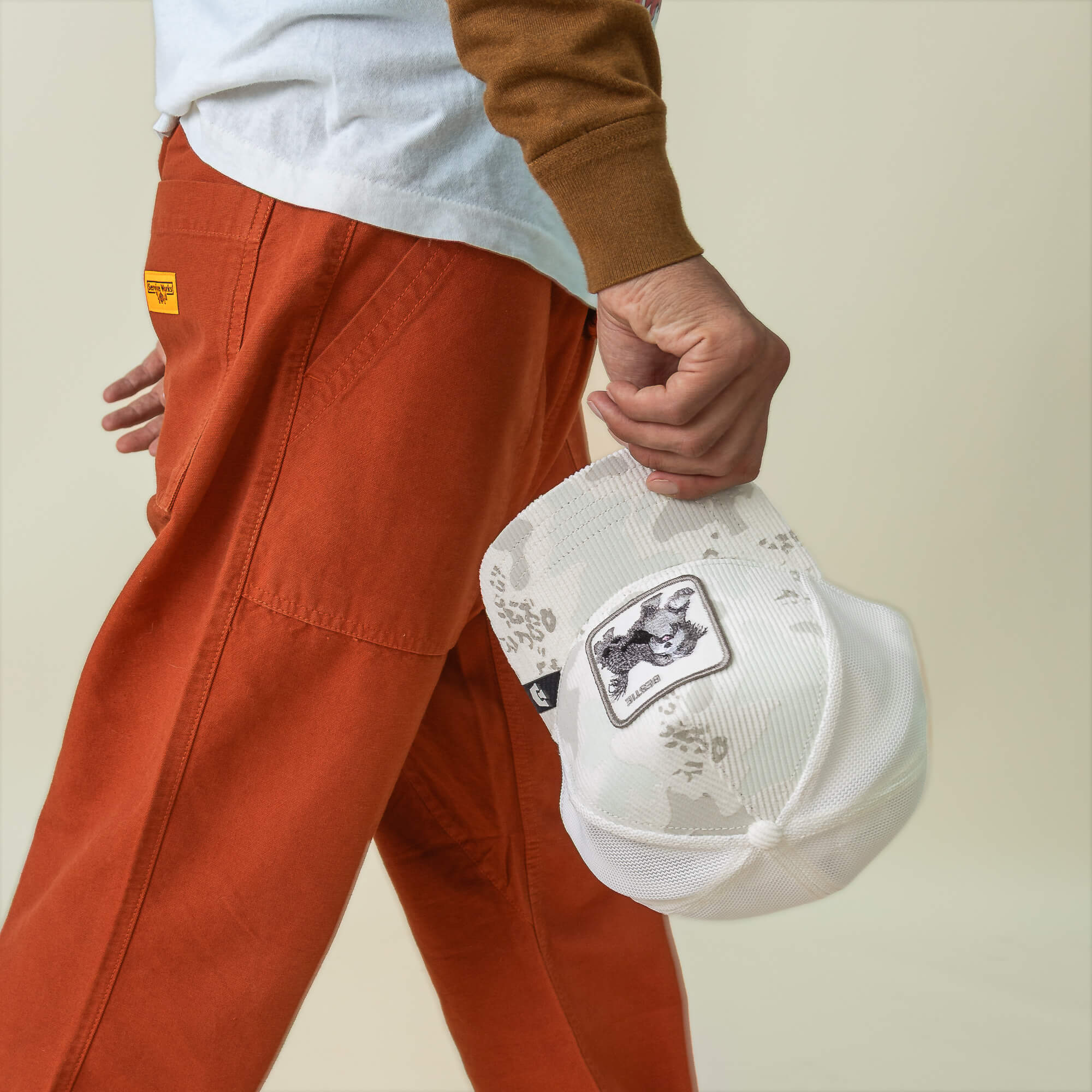 A person in orange pants and a white long-sleeve shirt holds the Goorin Bros. Bestie Camo corduroy mesh cap with a shaggy pup animal patch.