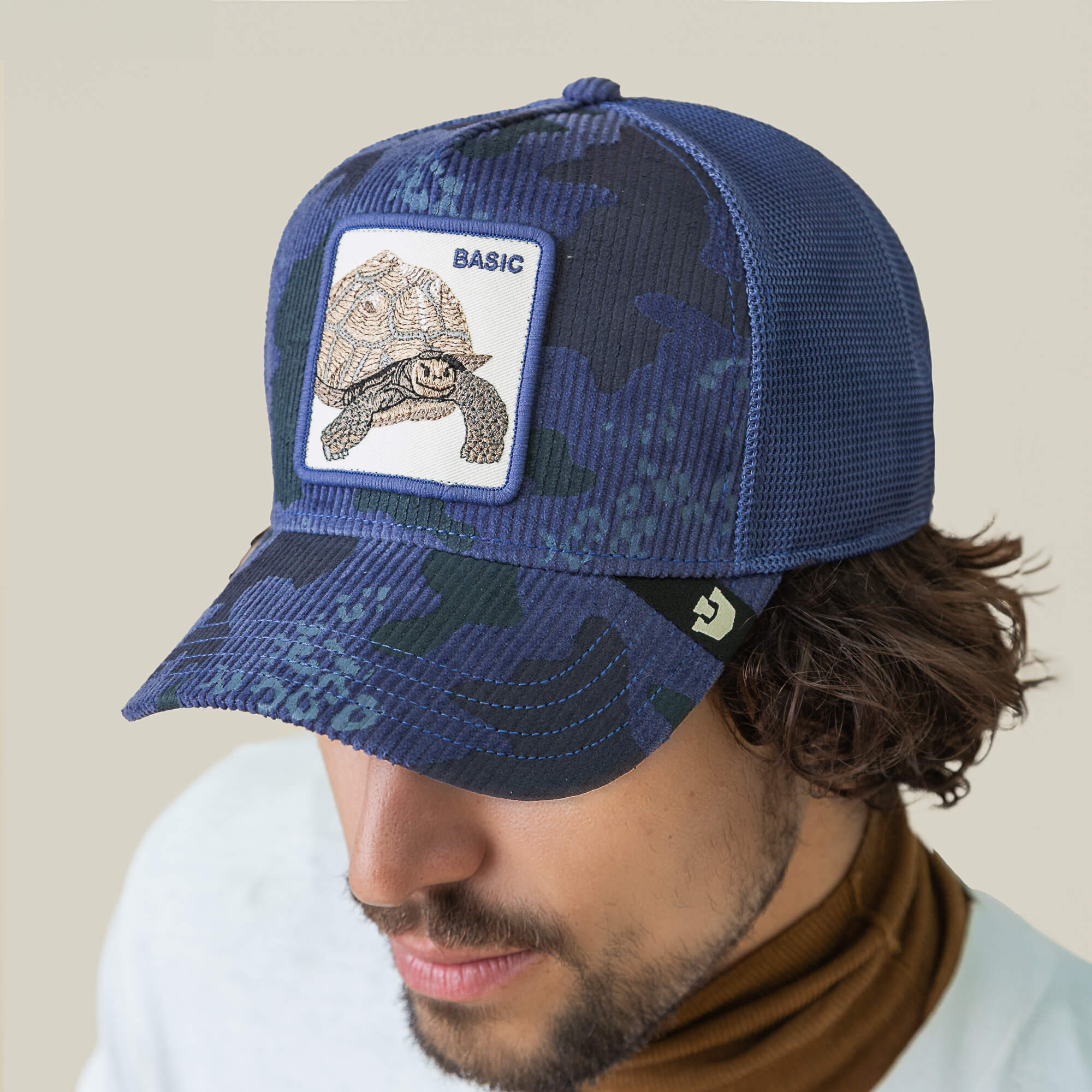 A man wears the Goorin Bros. Turtle Camo hat, featuring a blue camo corduroy front with a turtle patch labeled "BASIC" and a breathable mesh back.