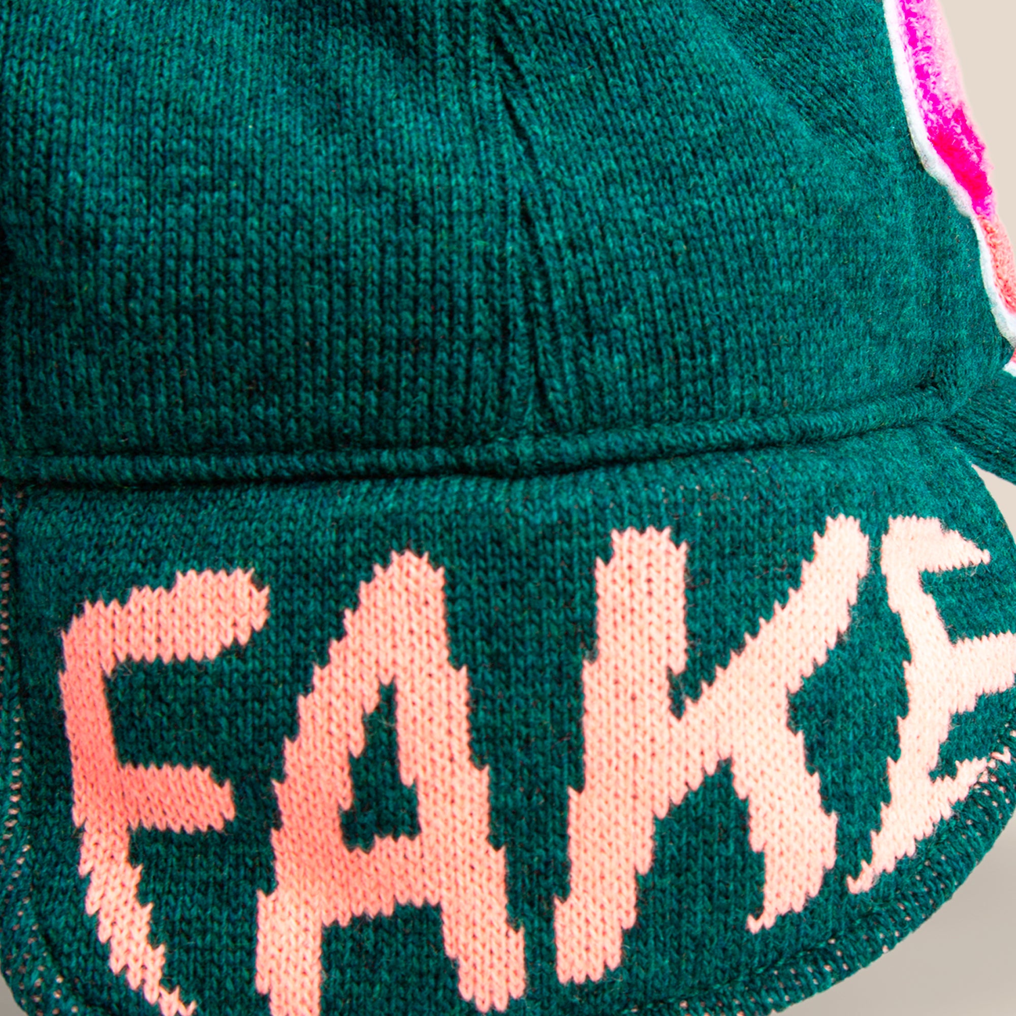 A close-up of the Goorin Bros. "F is for Fake" green knit fabric with FAKE stitched in large pink letters—a bold accessory for cold weather.