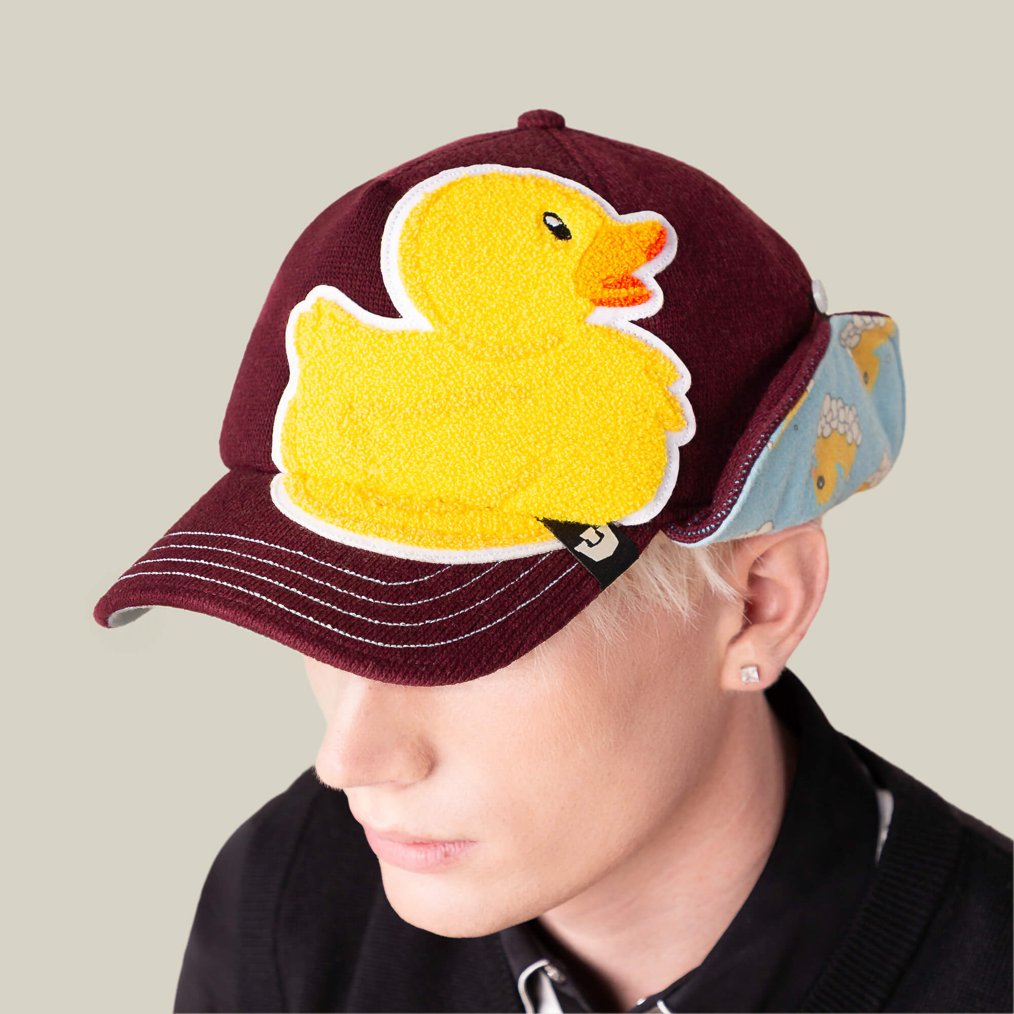 A person wearing the Goorin Bros. "F is for Duck" trucker hat, featuring a burgundy ear-flap design with a large yellow duck patch on the front and side fabric with duck patterns.