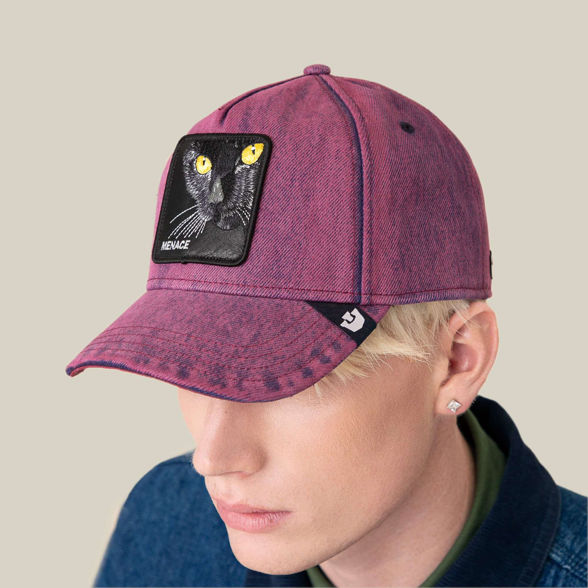 A person with light blonde hair wears the Goorin Bros. Rail Bird maroon cap, featuring a black cat patch labeled MENACE, radiating troublemaker energy against a plain beige background.