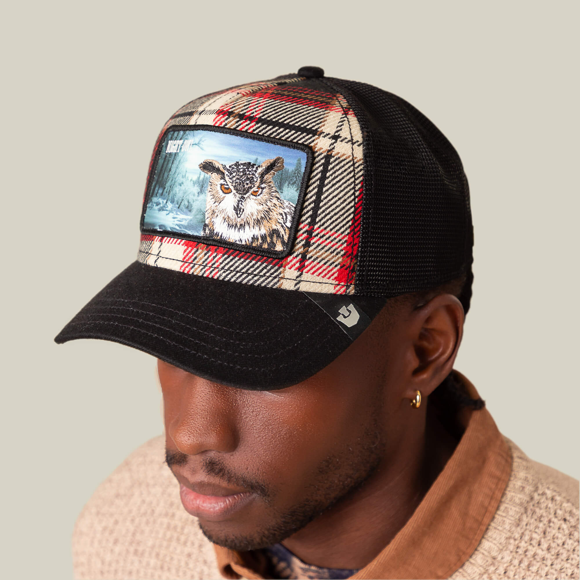 A man wears the Goorin Bros. "Cabin Fever" black mesh trucker cap with an embroidered owl patch, paired with a flannel jacket, against a plain light background.