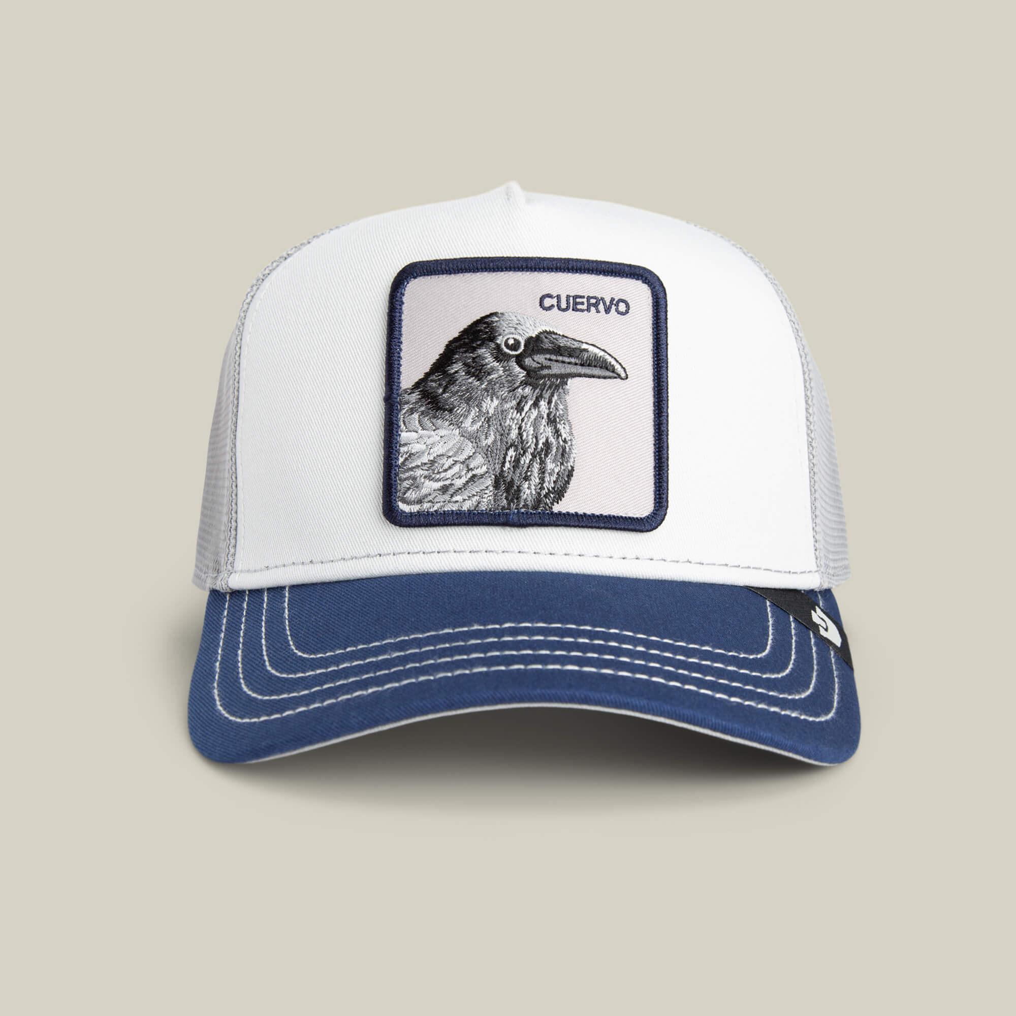 The Goorin Bros. Tricolor Raven hat is a white mesh trucker cap with a navy bill, featuring a raven patch and "CUERVO" above it, shown on a plain beige background.