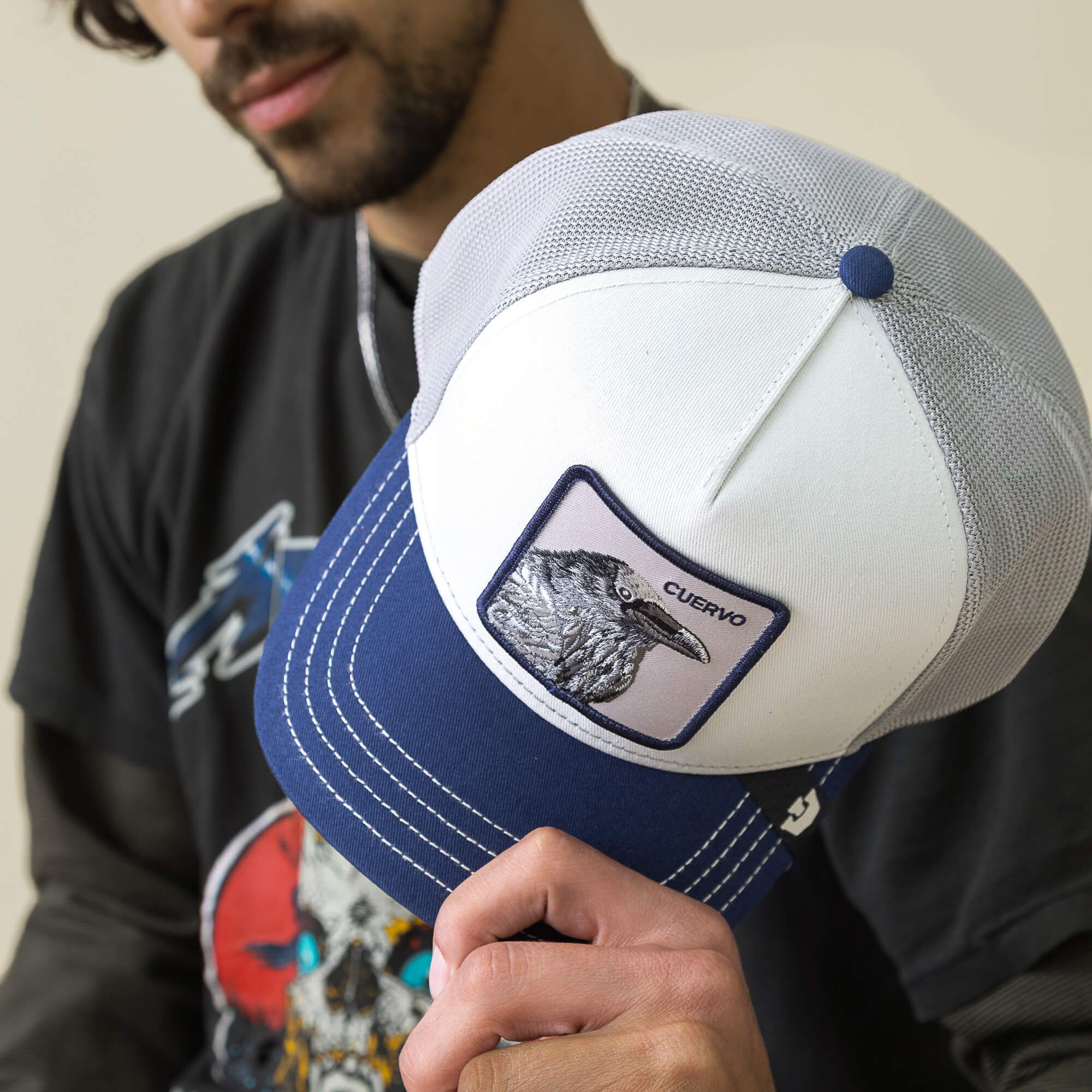 A person holds a Goorin Bros. Tricolor Raven hat—a white and blue mesh trucker cap with a patch showing a black and white raven illustration and the word “CUERVO.”.