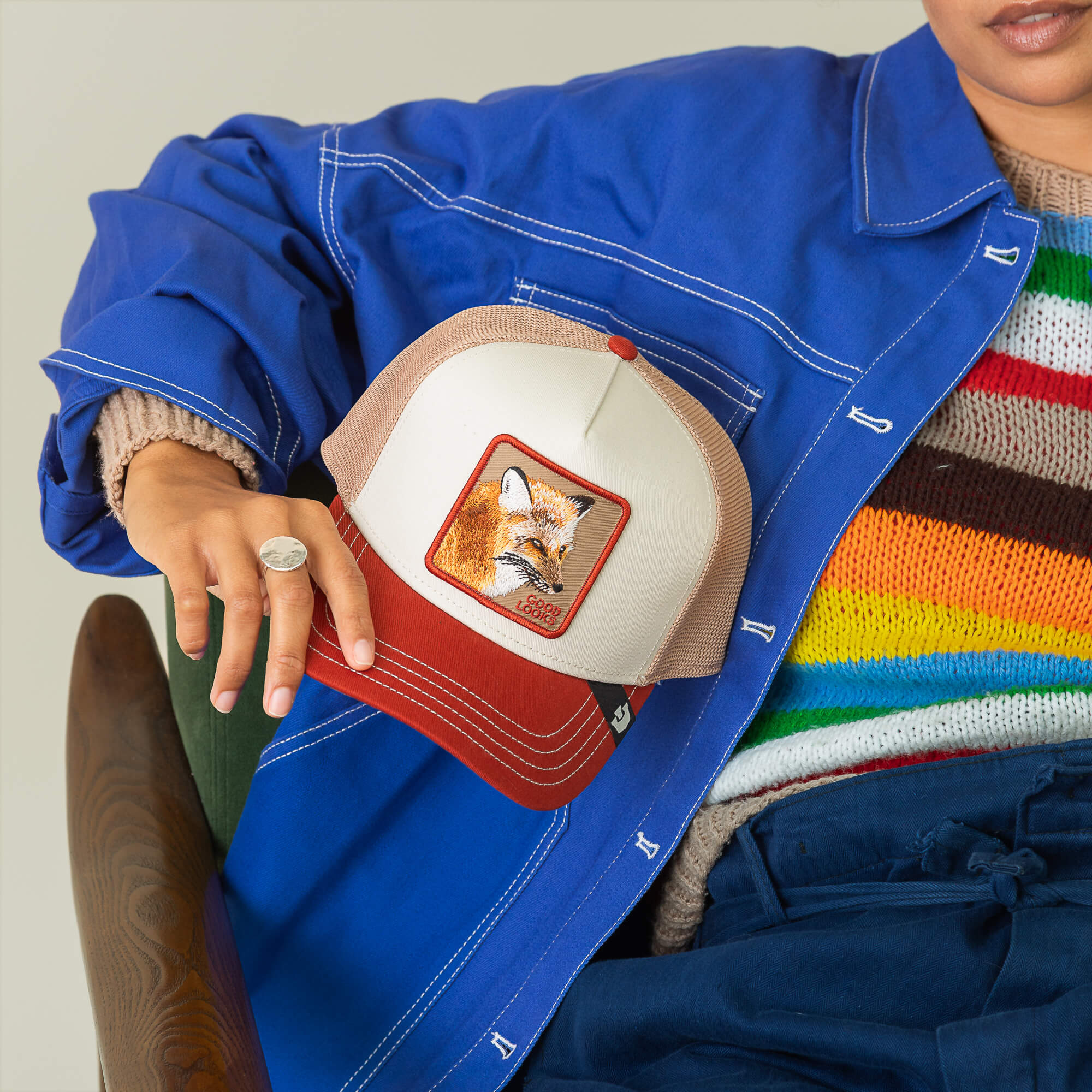 A person in a blue jacket and colorful striped sweater holds Goorin Bros.’ Tricolor Fox trucker hat, featuring a red and beige design with a bold fox patch on the front.