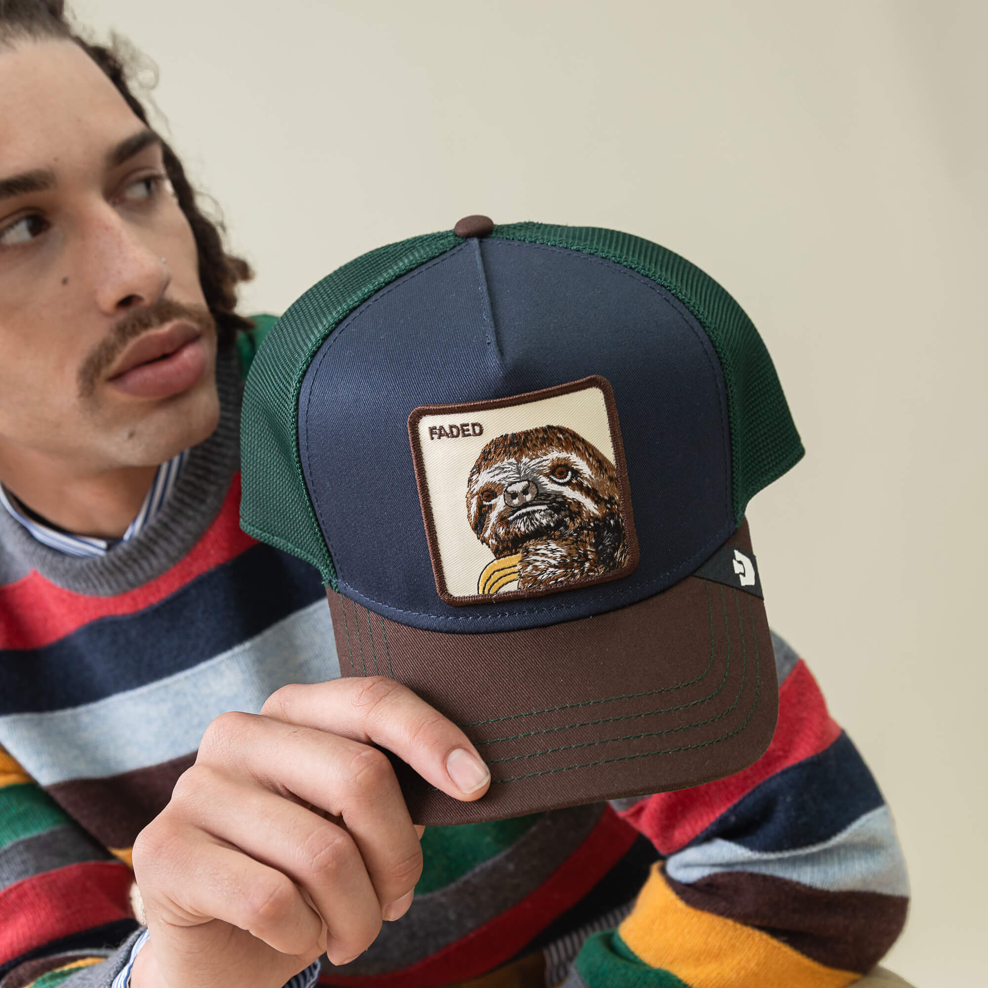 Someone in a striped sweater holds a Goorin Bros. Tricolor Sloth hat—a trucker style featuring a sloth patch and the word "FADED.