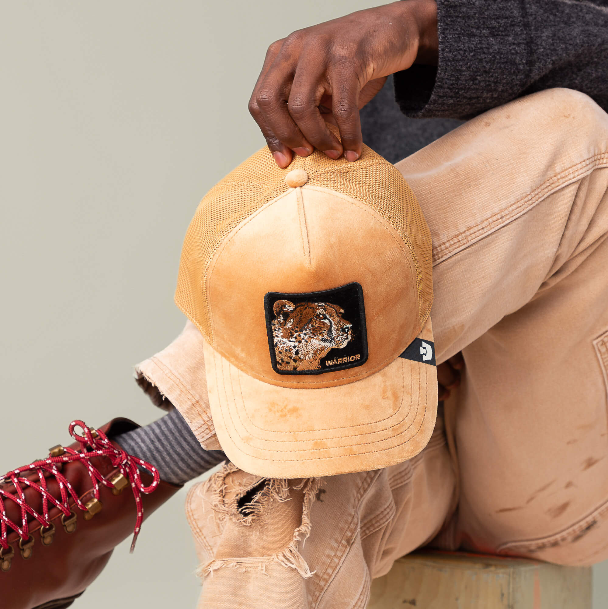 A person in tan pants and red boots holds the Goorin Bros. Velour Cheetah tan mesh baseball cap featuring a jaguar face patch and the word "WARRIOR" embroidered on it.