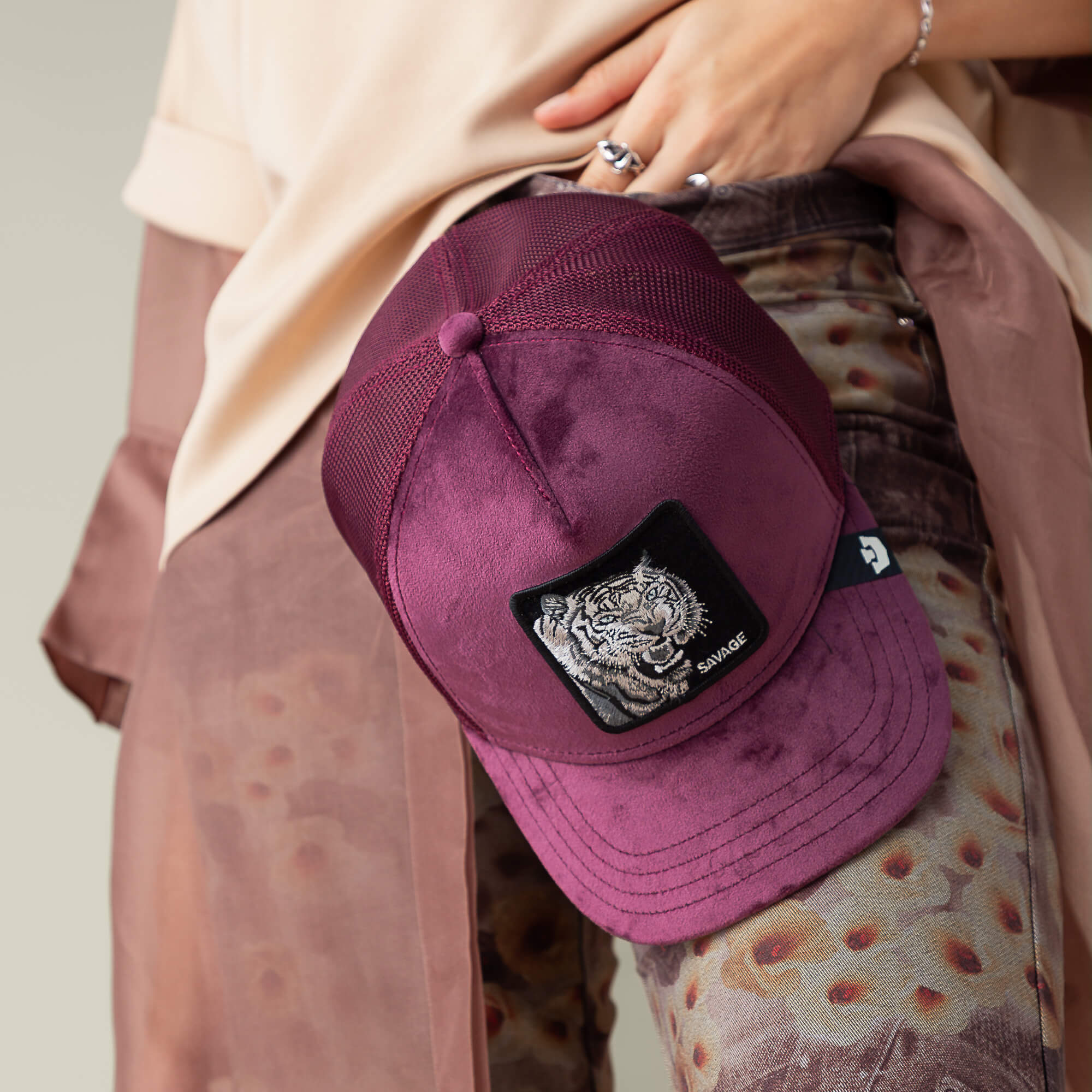 A person holds the Velour Tiger baseball cap by Goorin Bros.—a luxury maroon hat with a black patch showing a white tiger and "SAVAGE"—resting against deep wine-toned patterned pants.