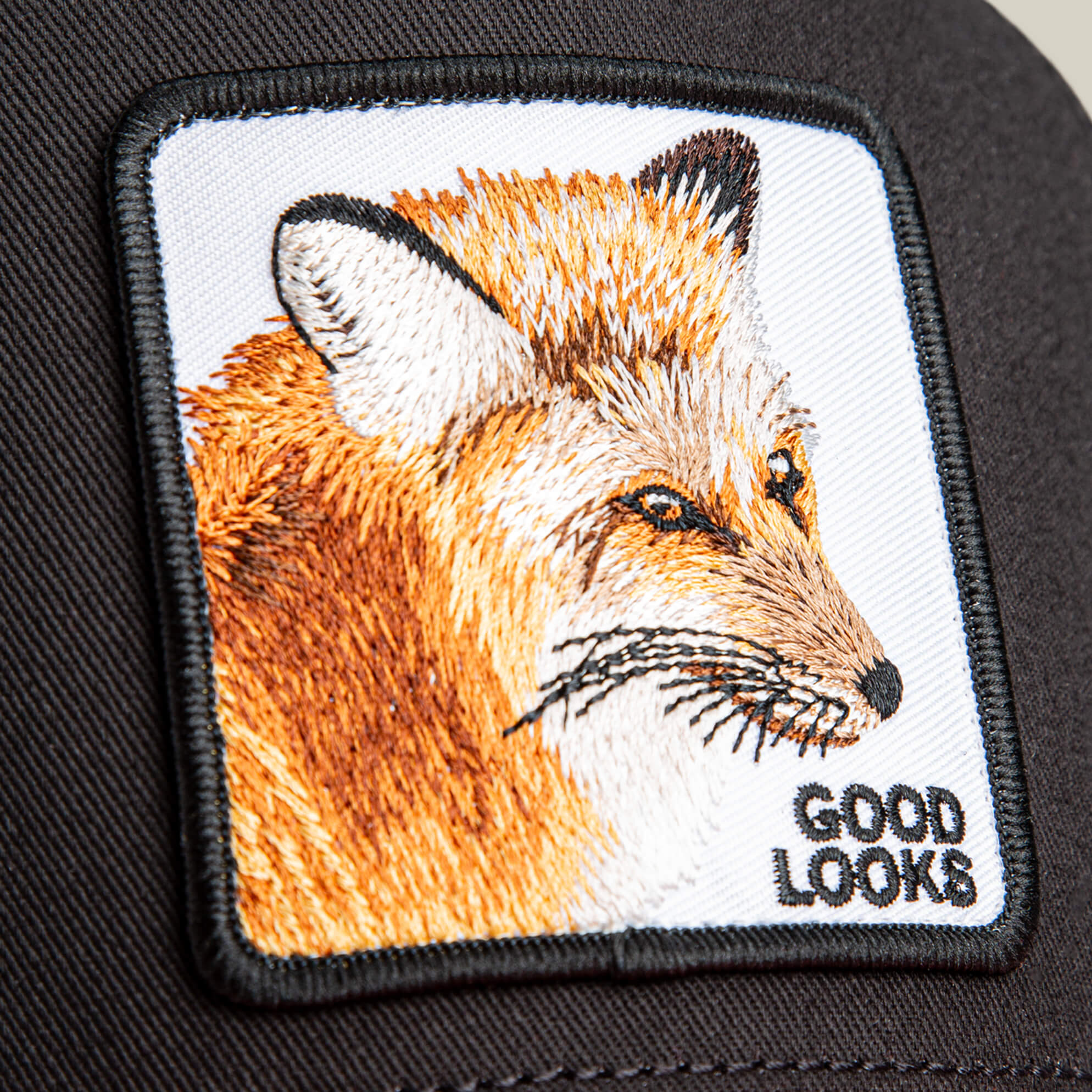 Close-up of the Goorin Bros. "Good Looks Fox" trucker hat, featuring an embroidered fox head patch with "GOOD LOOKS" in black text at the lower right corner.