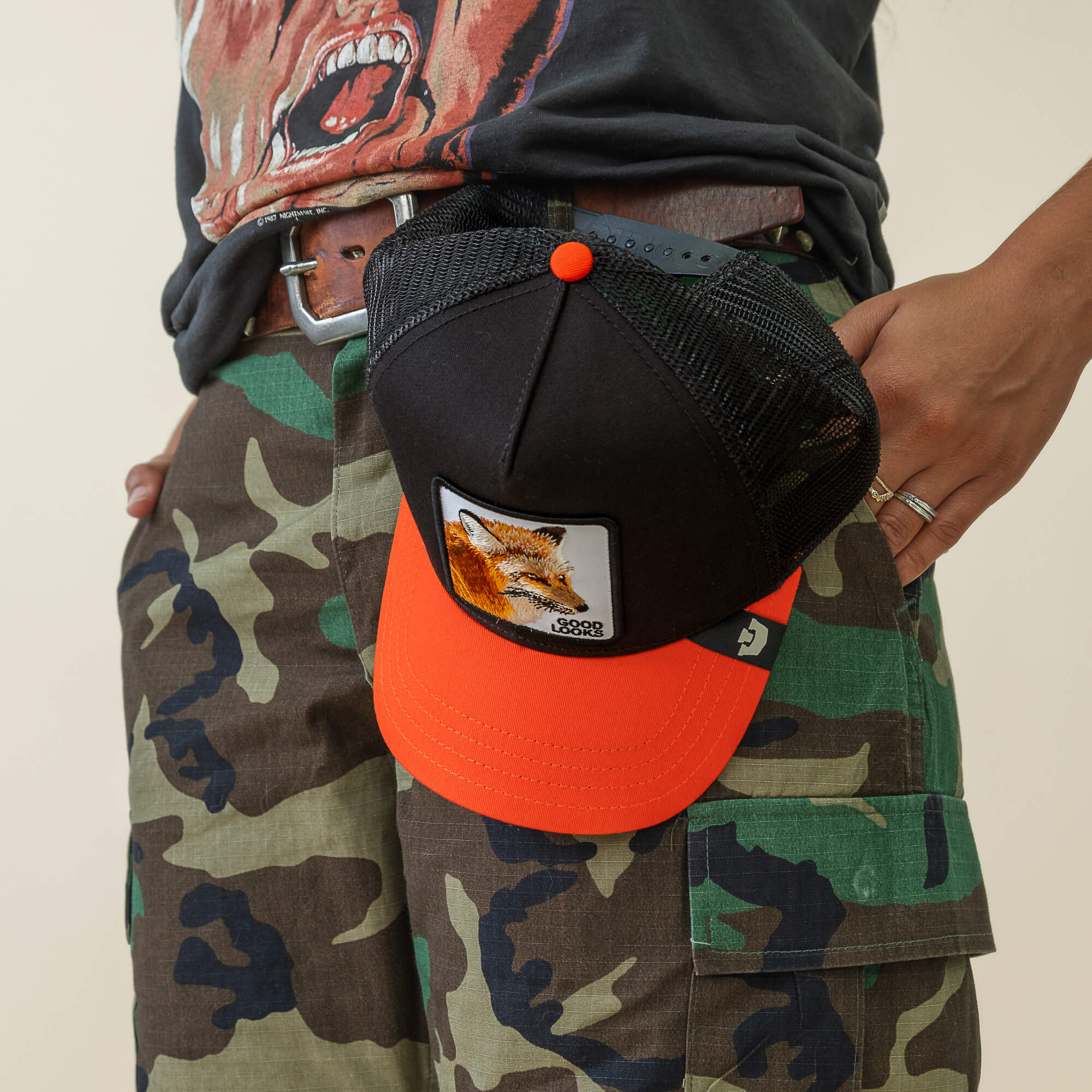 Wearing camouflage cargo shorts and a graphic tee, someone hooks the Goorin Bros. Good Looks Fox trucker hat—featuring an orange and black fox patch—to their belt, merging farmyard chic with effortless style.