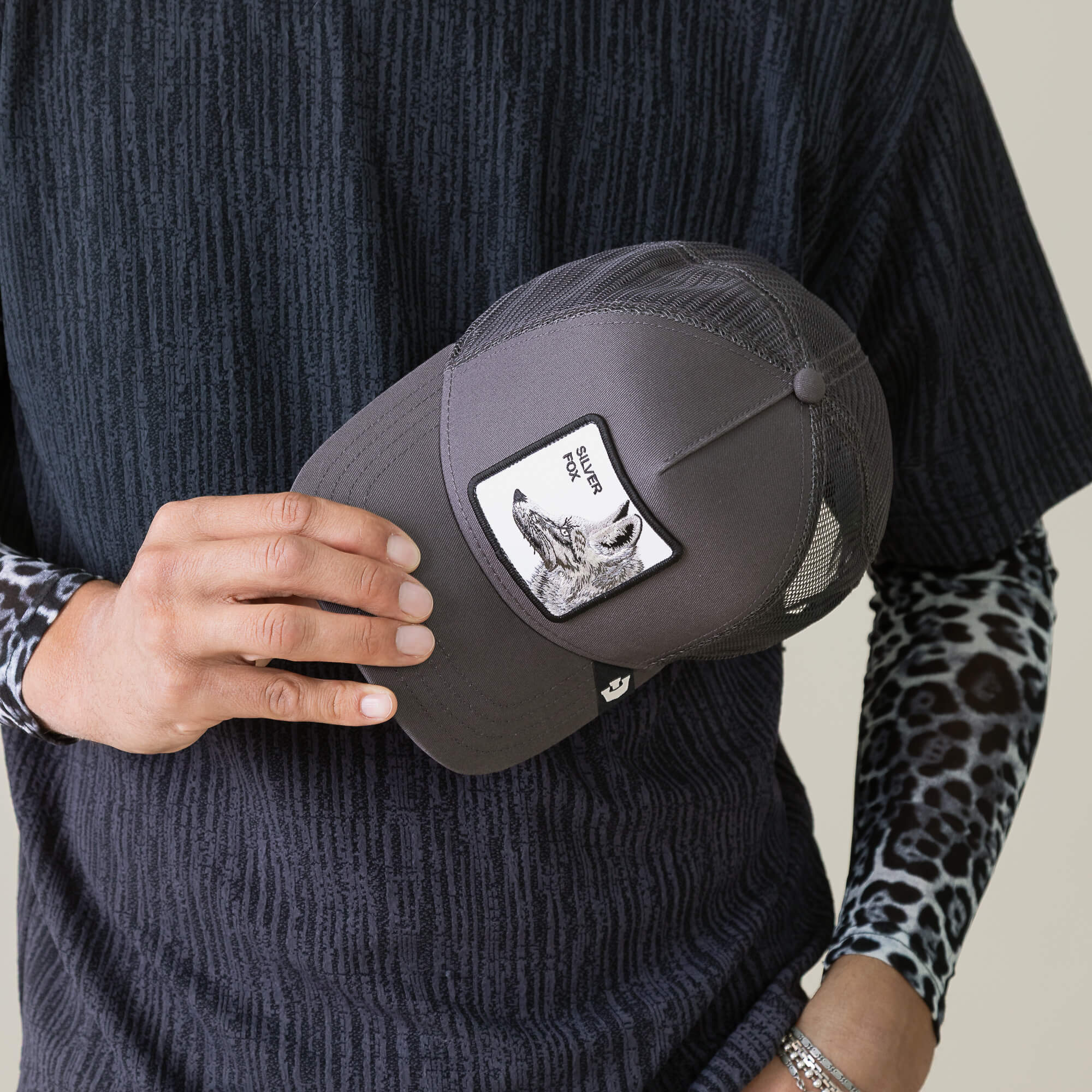 A person in a dark textured shirt with leopard print sleeves holds a stylish Goorin Bros. cap, the “Silver Foxxx,” featuring a wolf patch and the words SILVER WOLF, echoing the refined allure of the Silver Fox.
