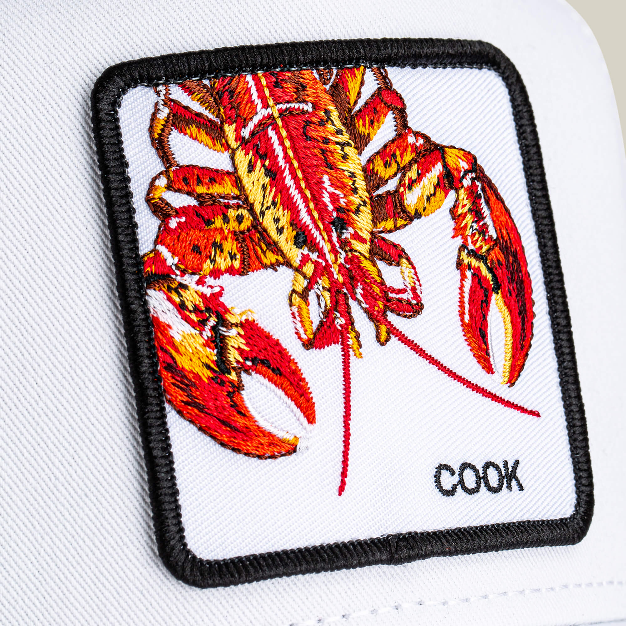 Close-up of a white patch with an embroidered red lobster and "COOK" stitched at the corner, featured on the Lemme Cook trucker hat by Goorin Bros., showcasing a stylish mesh back design.
