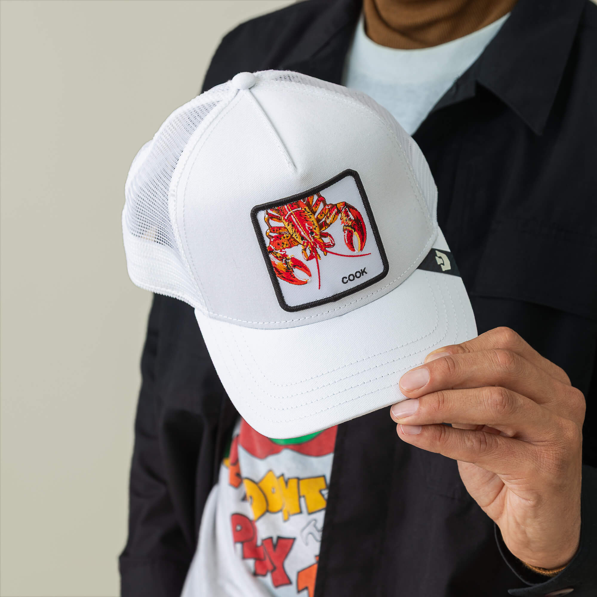 A person holds the Lemme Cook trucker cap by Goorin Bros., a white mesh-back baseball hat with a patch featuring a red lobster and the word "COOK" embroidered on it.