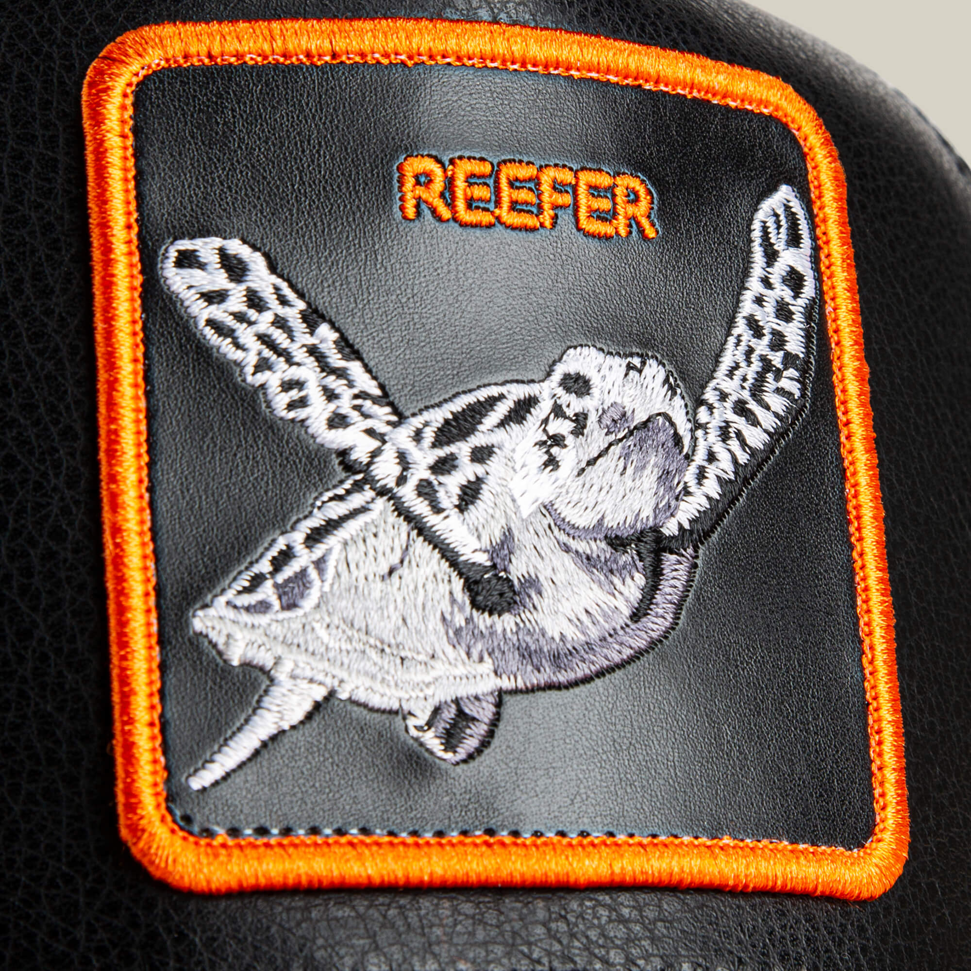 A rectangular orange-bordered patch with an embroidered sea turtle and "REEFER" is sewn onto black faux leather Courtside Reefer sneakers by Goorin Bros.