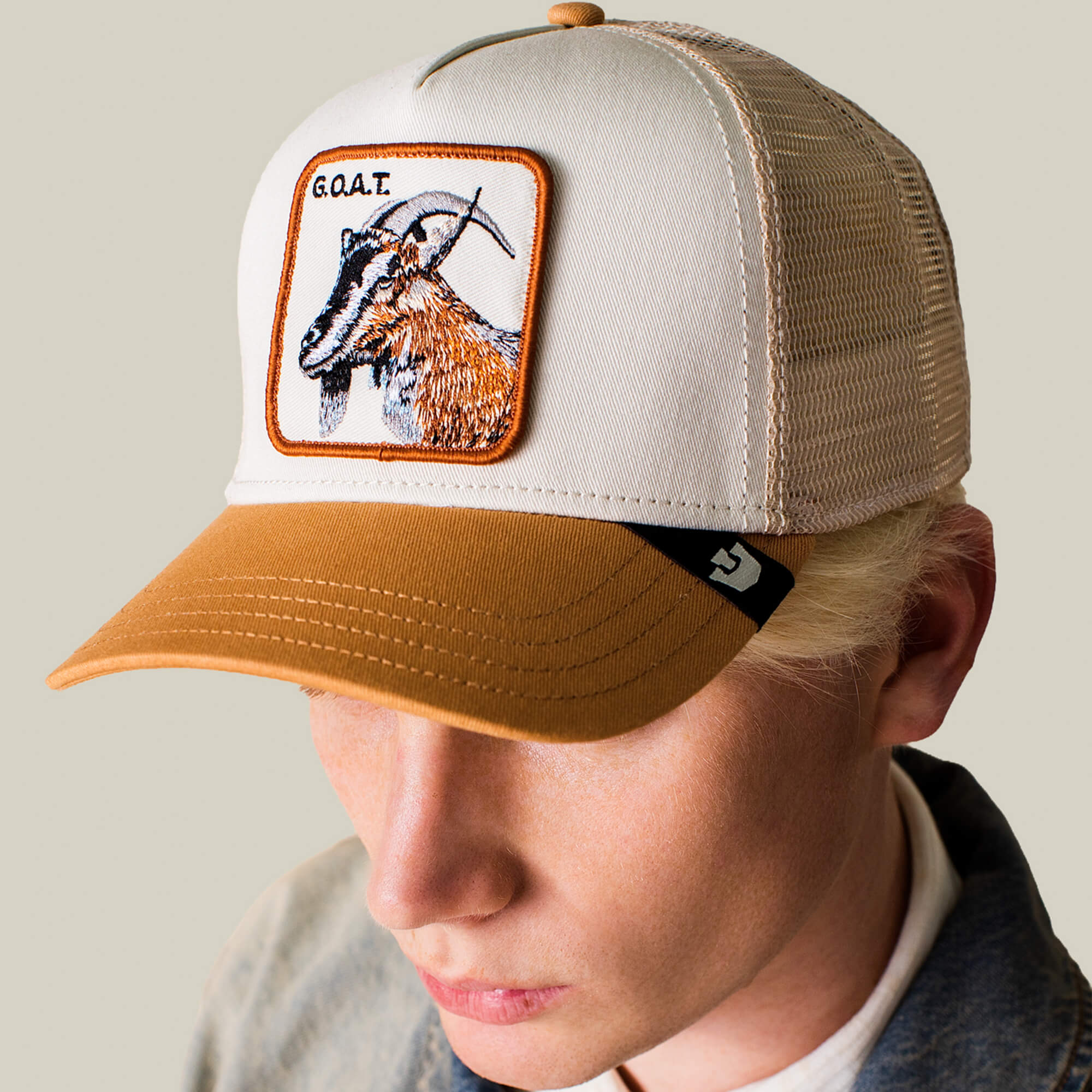 A person wears The G.O.A.T hat by Goorin Bros.—a beige and brown cap in neutral tones, featuring a bold front patch with a goat and the text "G.O.A.T.