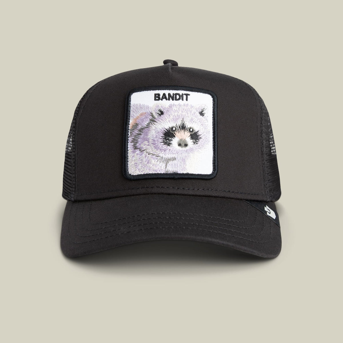 The Goorin Bros. Bandit Trucker is a black mesh trucker hat featuring an embroidered patch of a raccoon with the word Bandit on a white background, displayed against a neutral studio backdrop.