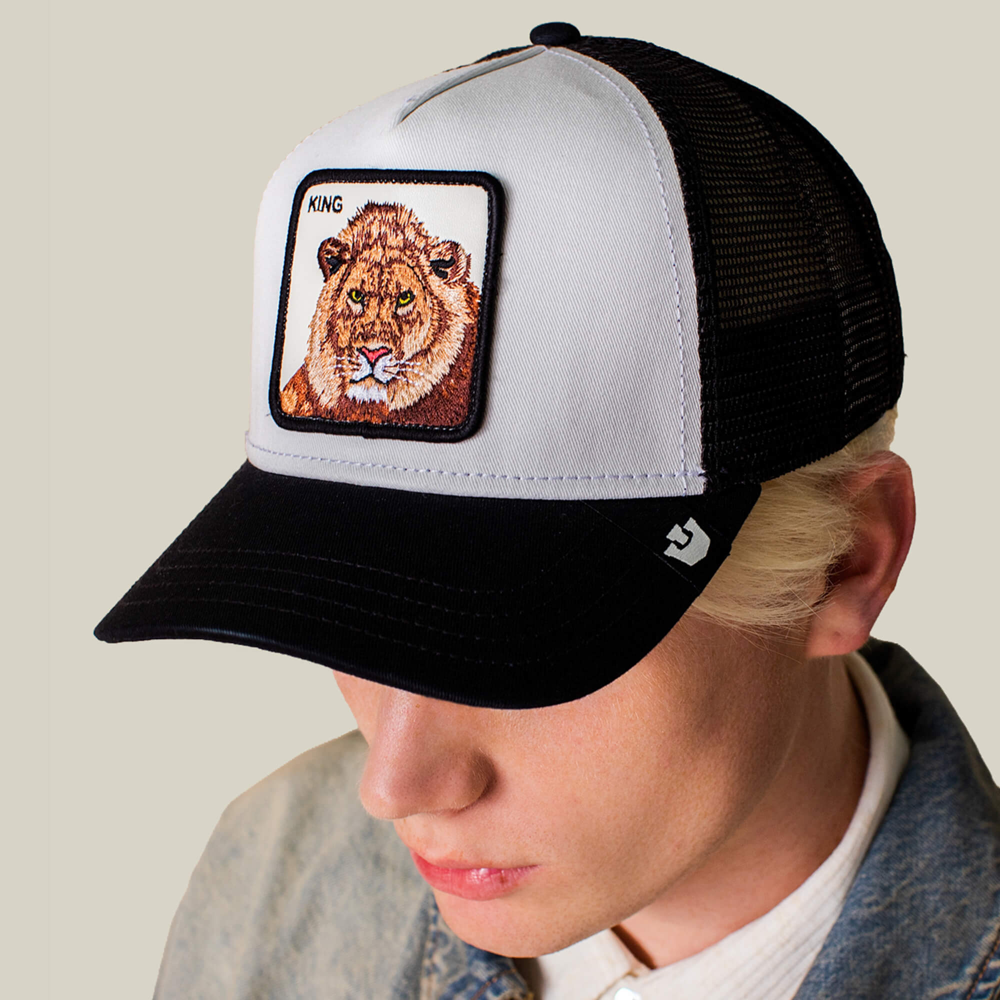 Wearing the Goorin Bros. King Trucker hat, featuring a black and white design with a KING lion patch, paired with a light denim jacket, this person exudes confident power dressing while looking downward.