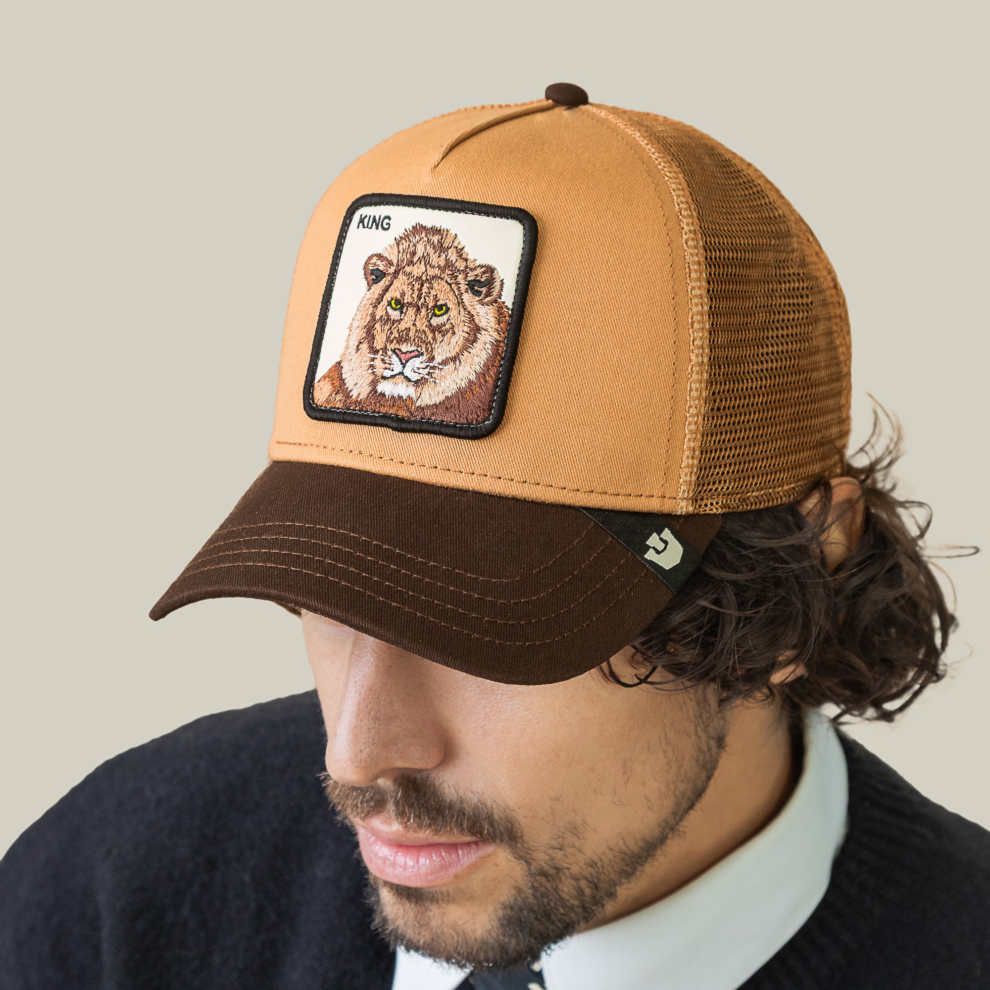 A man wears the Goorin Bros. King Trucker, a brown and tan mesh hat featuring a KING lion patch on the front, and looks down against a plain beige background.