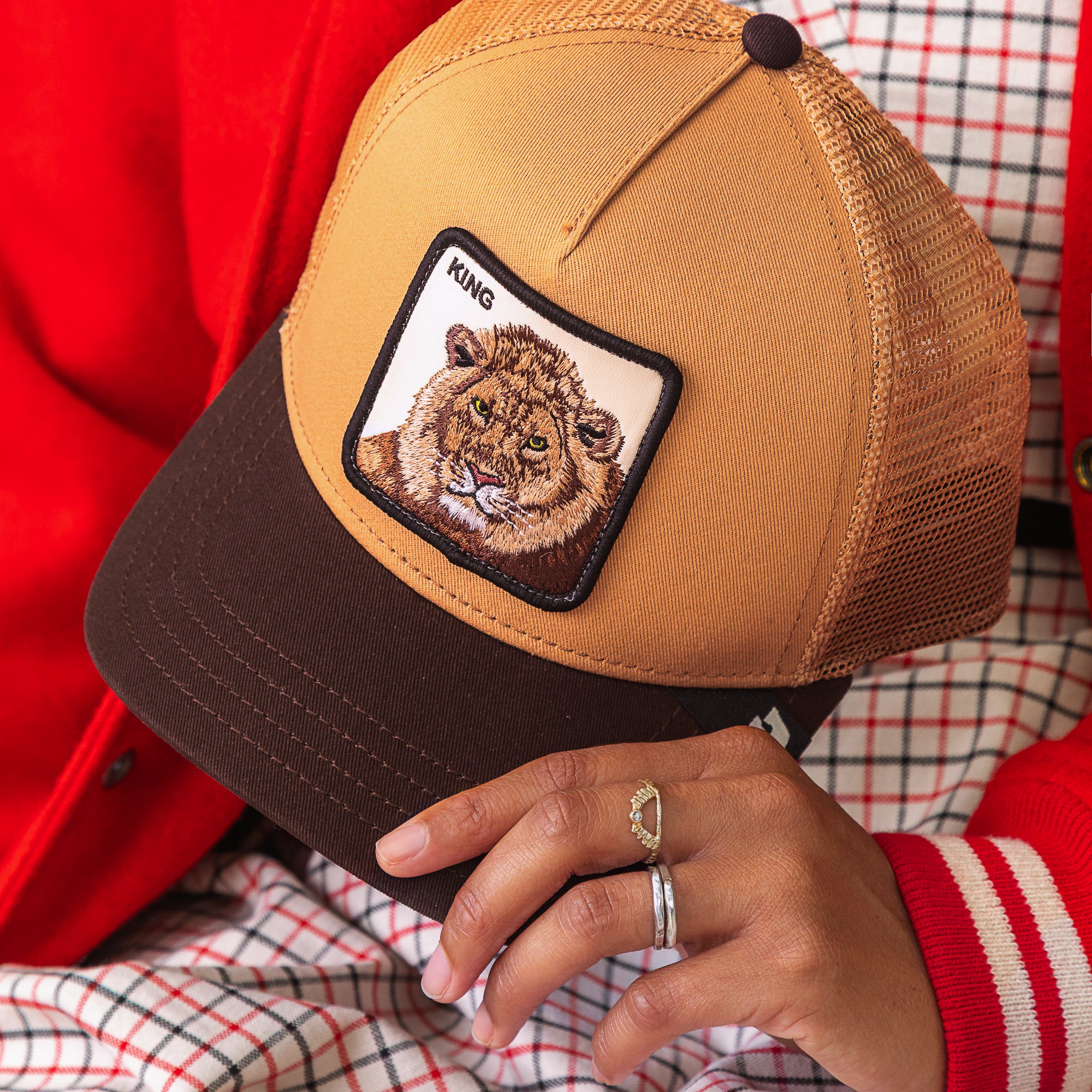 Wearing rings and dressed in power style with a red jacket and checkered clothes, they confidently hold the Goorin Bros. King Trucker cap—a brown and tan mesh hat featuring a bold lion patch labeled KING.
