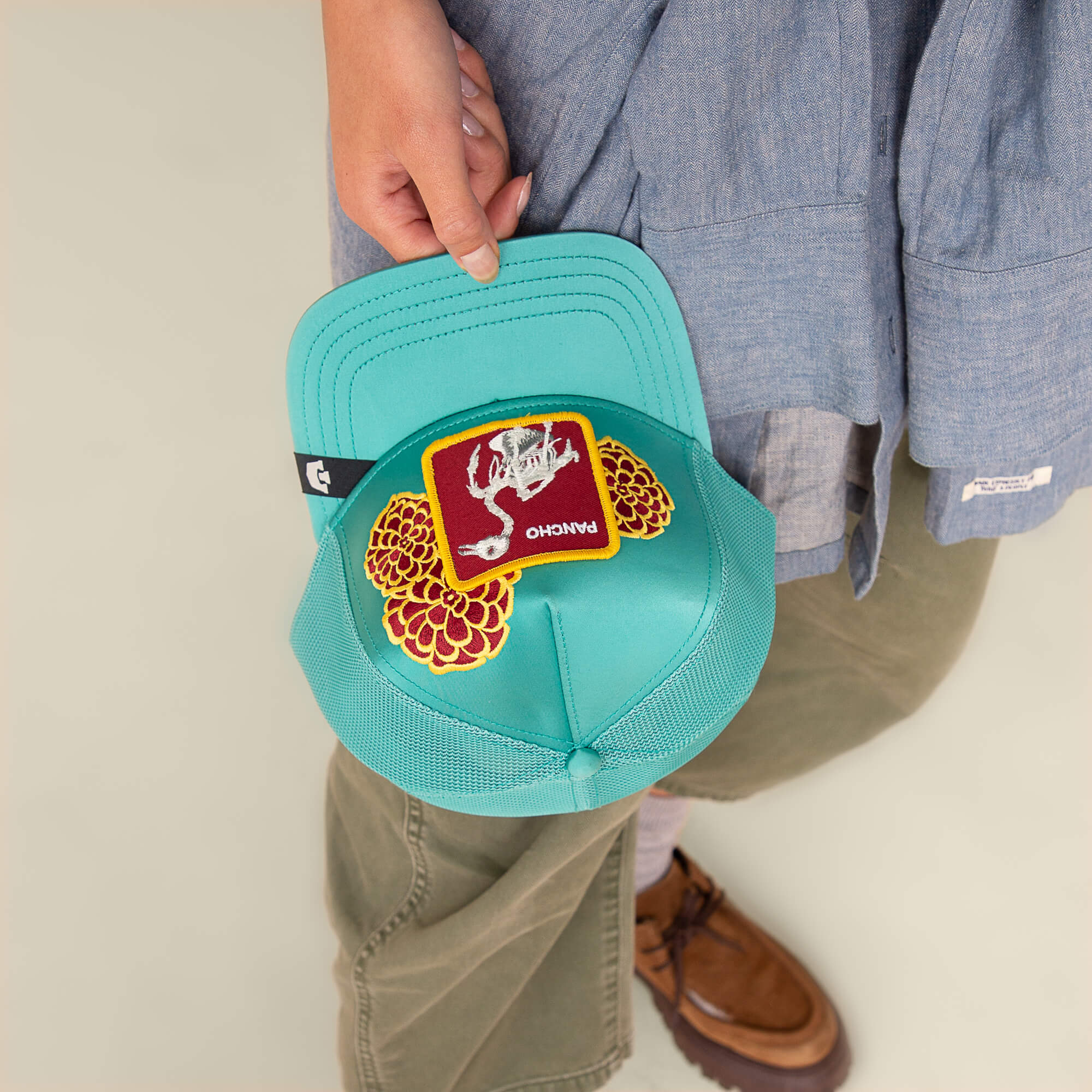 Someone holds the Goorin Bros. "Dia De Pancho" teal mesh cap, featuring a stitched bone red Pancho patch and two yellow-red patches, embodying the rebel spirit of Día de los Muertos against a light background.