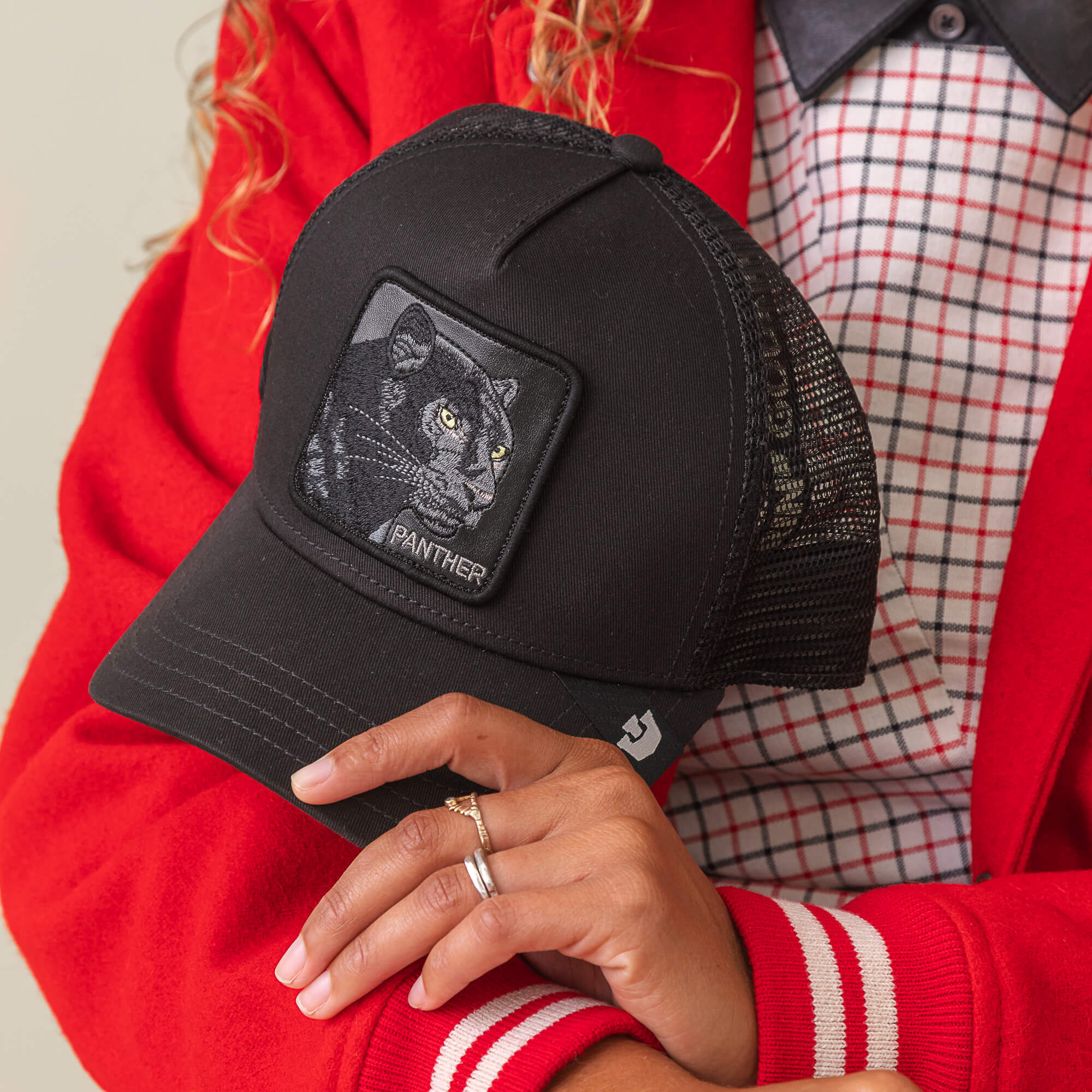 A person holds The Panther hat by Goorin Bros., featuring a black mesh cap and panther patch, paired with a red and white jacket over a checked shirt for a trendy look.