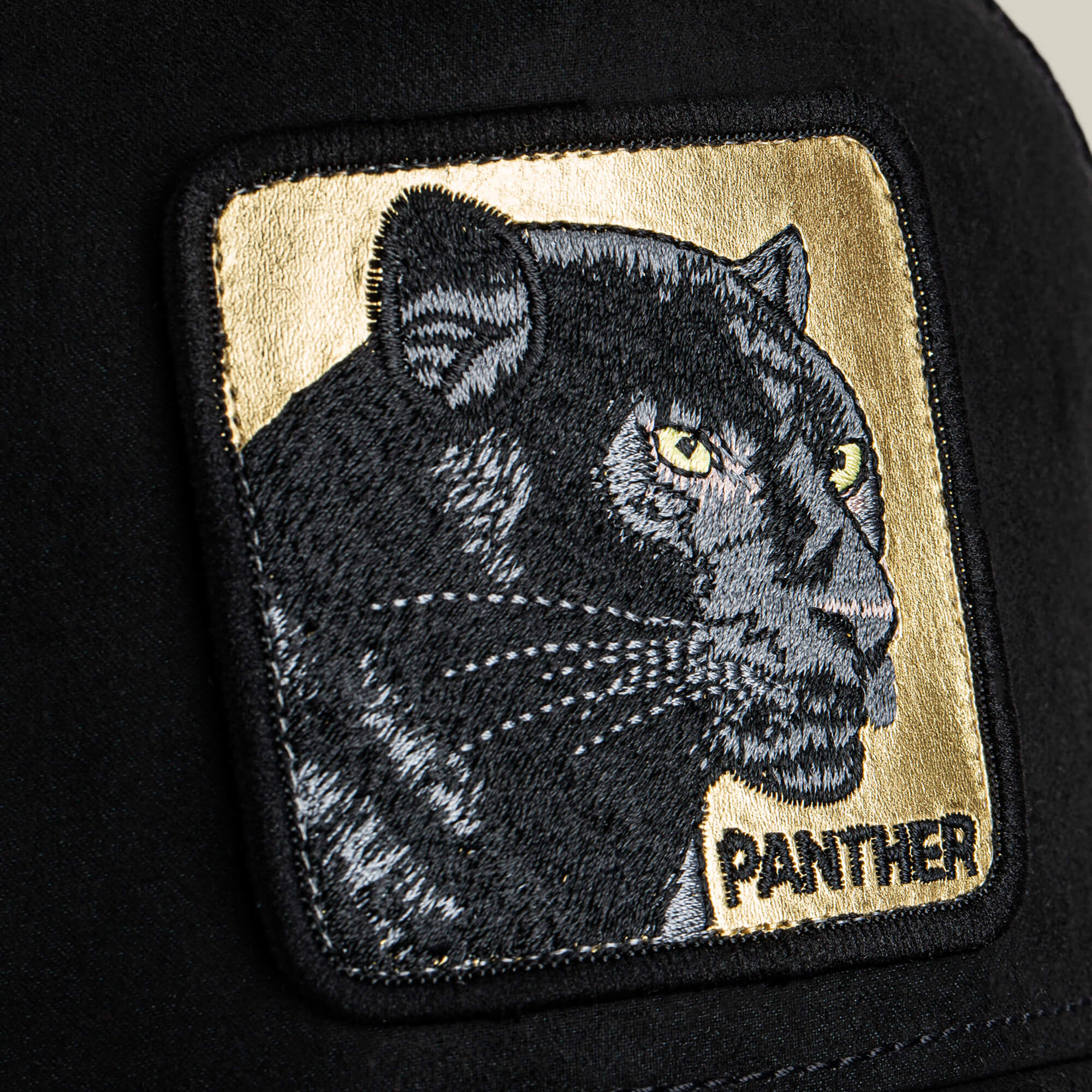 A close-up of Goorin Bros. Golden Panther patch, showing a black panther with yellow eyes and "PANTHER" on gold, sewn onto a stealth hat with a microsuede crown.