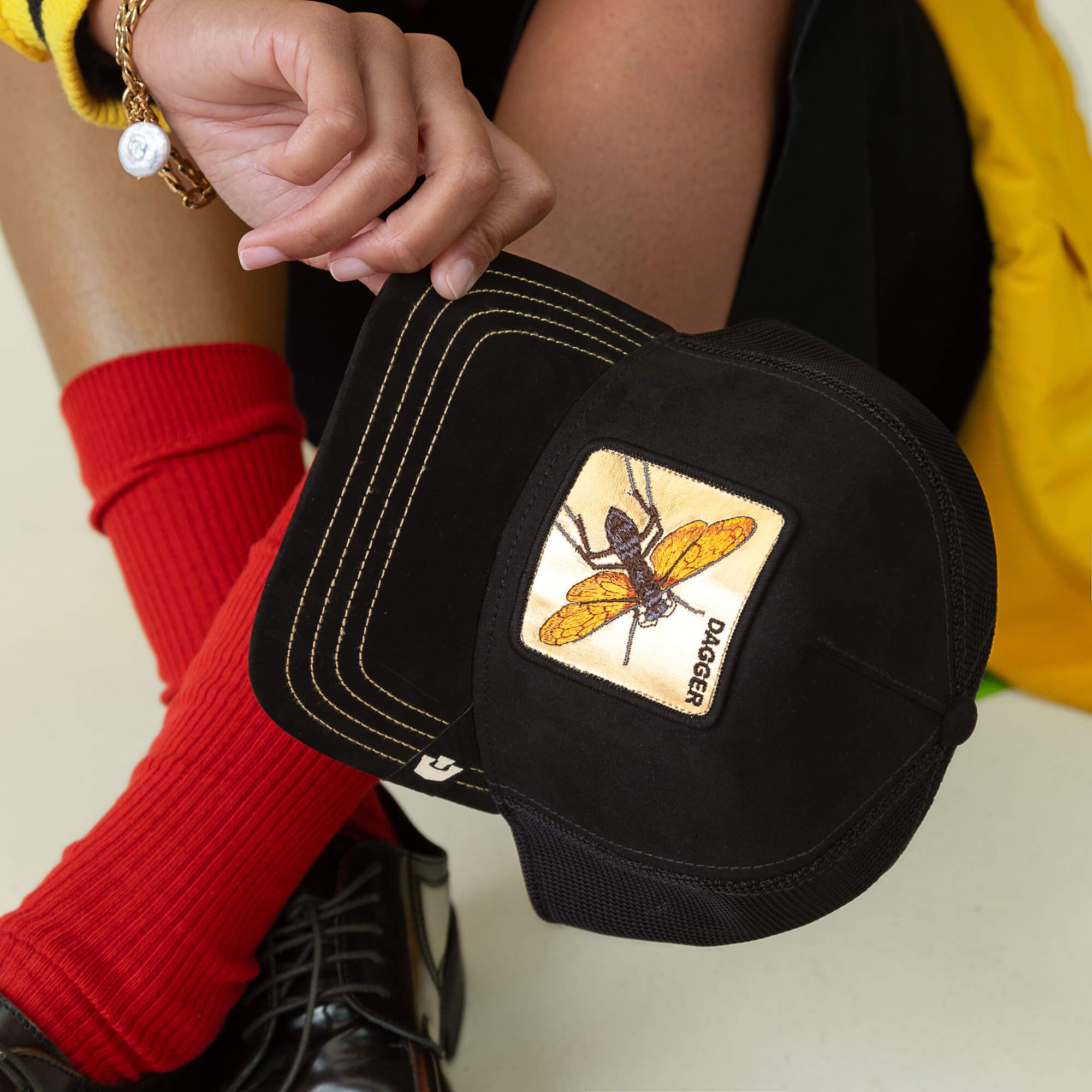 A person in red socks and black shoes holds the Goorin Bros. Golden Dagger cap, a sharp black hat with a yellow insect patch labeled "DAGGER.