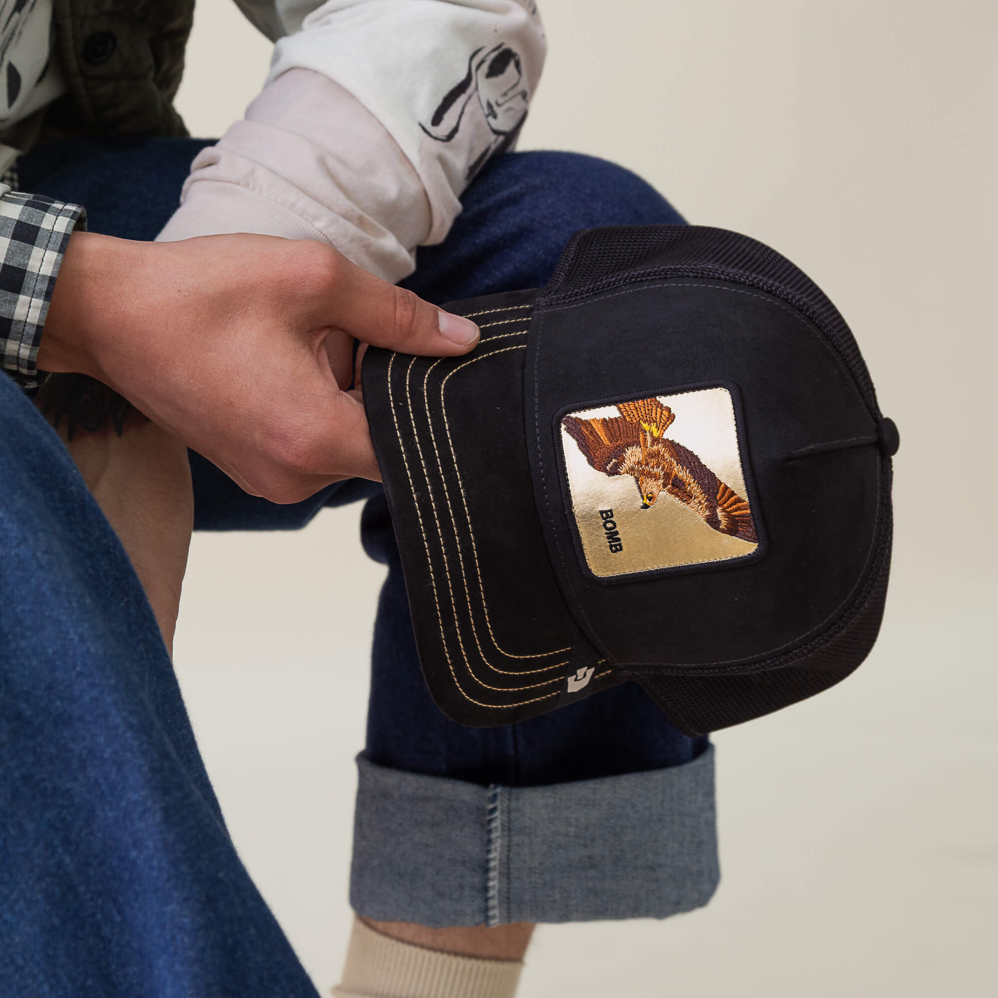 A person holds the Golden Bomb trucker hat by Goorin Bros., featuring a black design with an eagle patch and "BIRD" embroidery—only their hand and part of their outfit are visible.