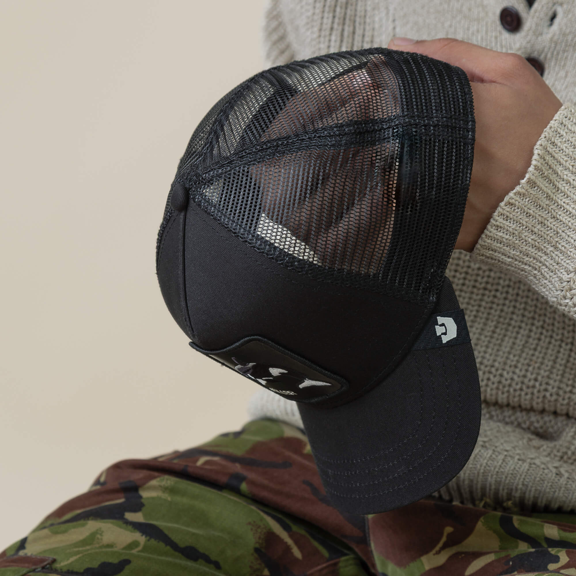 A person in a knit sweater and camo pants holds the Goorin Bros. Killer Trucker—a black mesh trucker hat with a front patch.