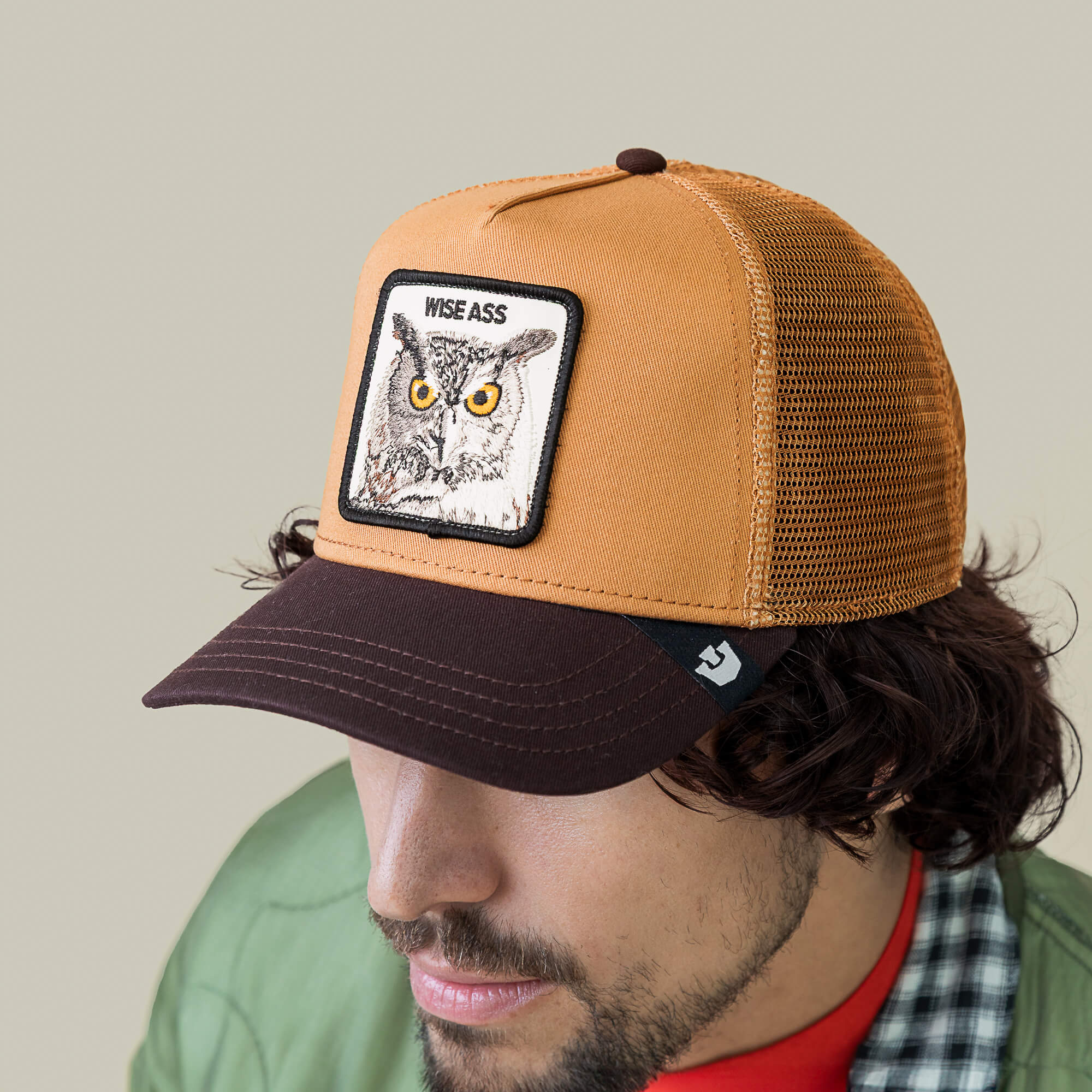 A man wearing a Goorin Bros. Wise Ass Trucker cap, featuring a brown and tan mesh design with an owl patch, along with a green jacket and red shirt.