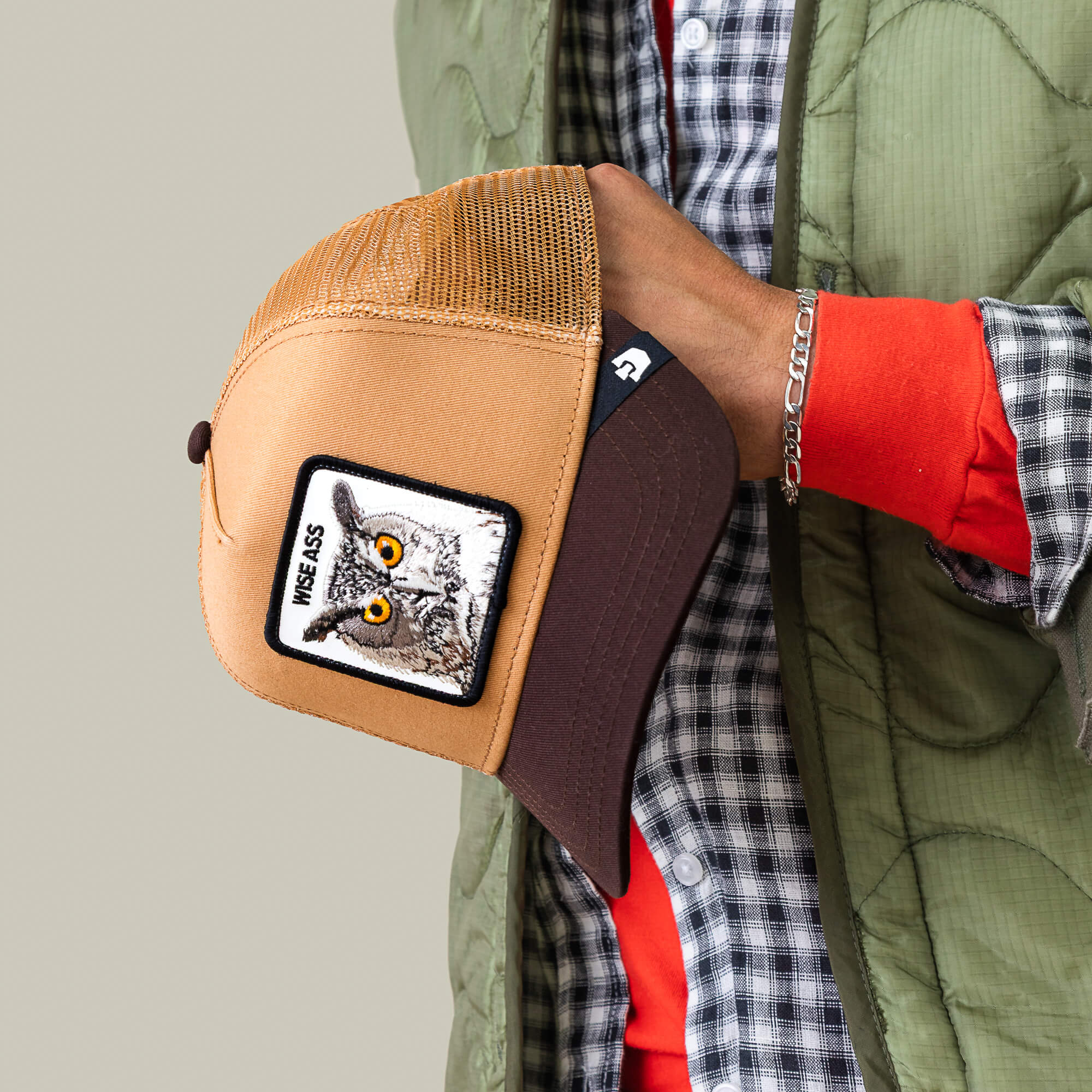 A person in a green quilted jacket holds the Goorin Bros. Wise Ass Trucker—a brown mesh cap with an owl patch and “WISE ASS” text.