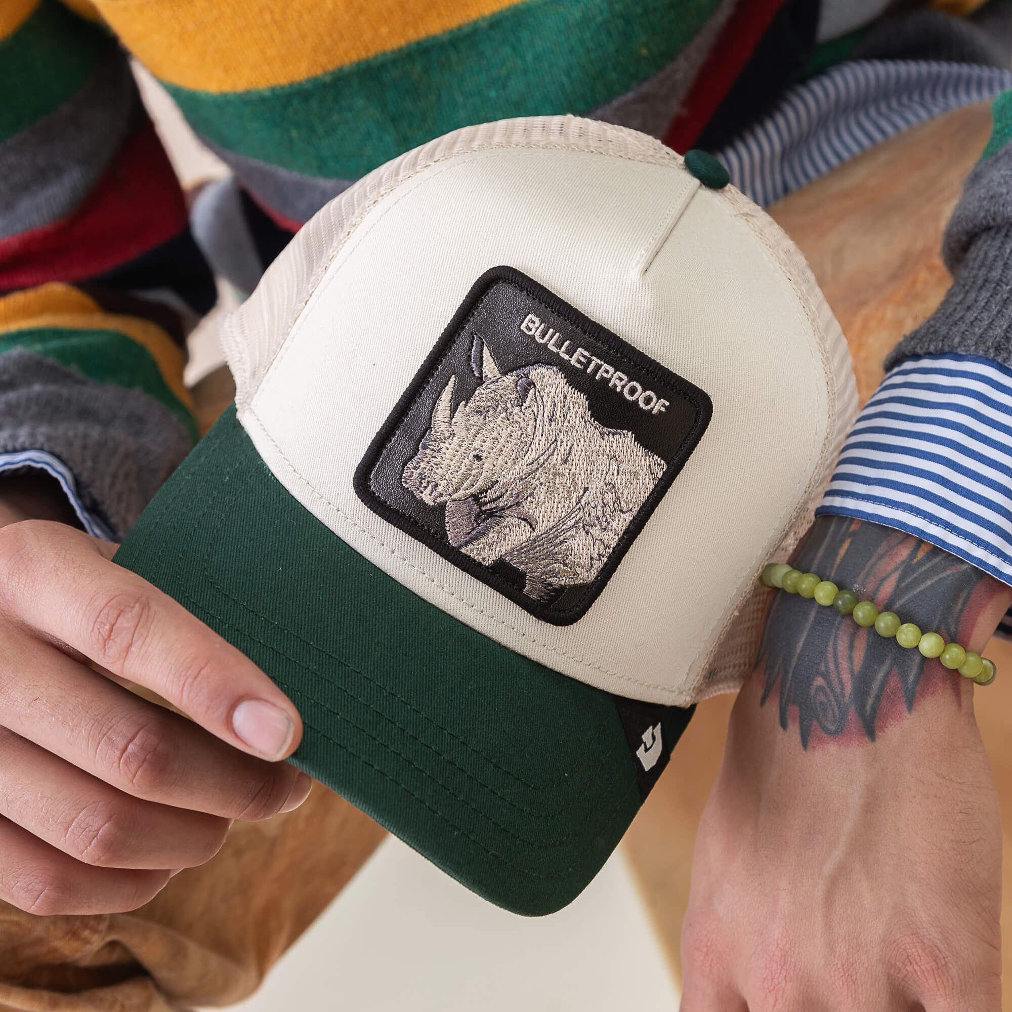 A person with a tattooed arm wears a beaded bracelet and striped sweater, holding the Goorin Bros. Bulletproof Trucker hat—beige and green with a rhino patch labeled "BULLETPROOF" in tribute to rhino armor.