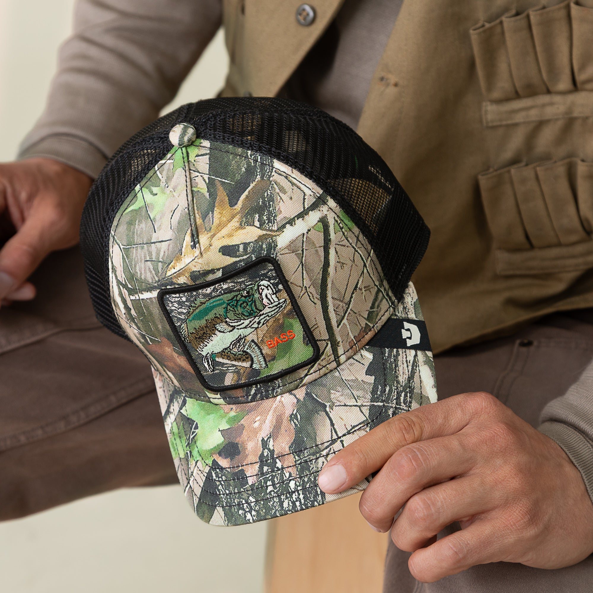 Person holding a Goorin Bros. Camouflage Bass trucker hat with mesh back, blending fishing fashion and outdoor style, featuring a bass patch and bold design.