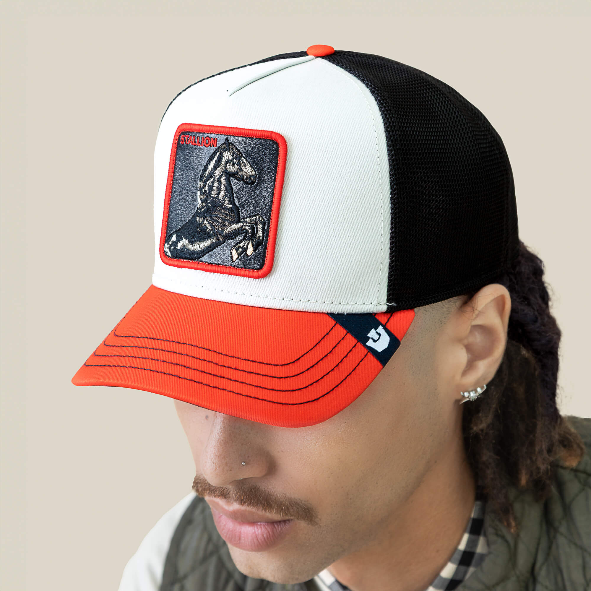 Someone wearing the Goorin Bros. Tricolor Stallion hat, a black mesh trucker with a bright orange visor and a patch that displays an embroidered stallion and the word "Stallion.